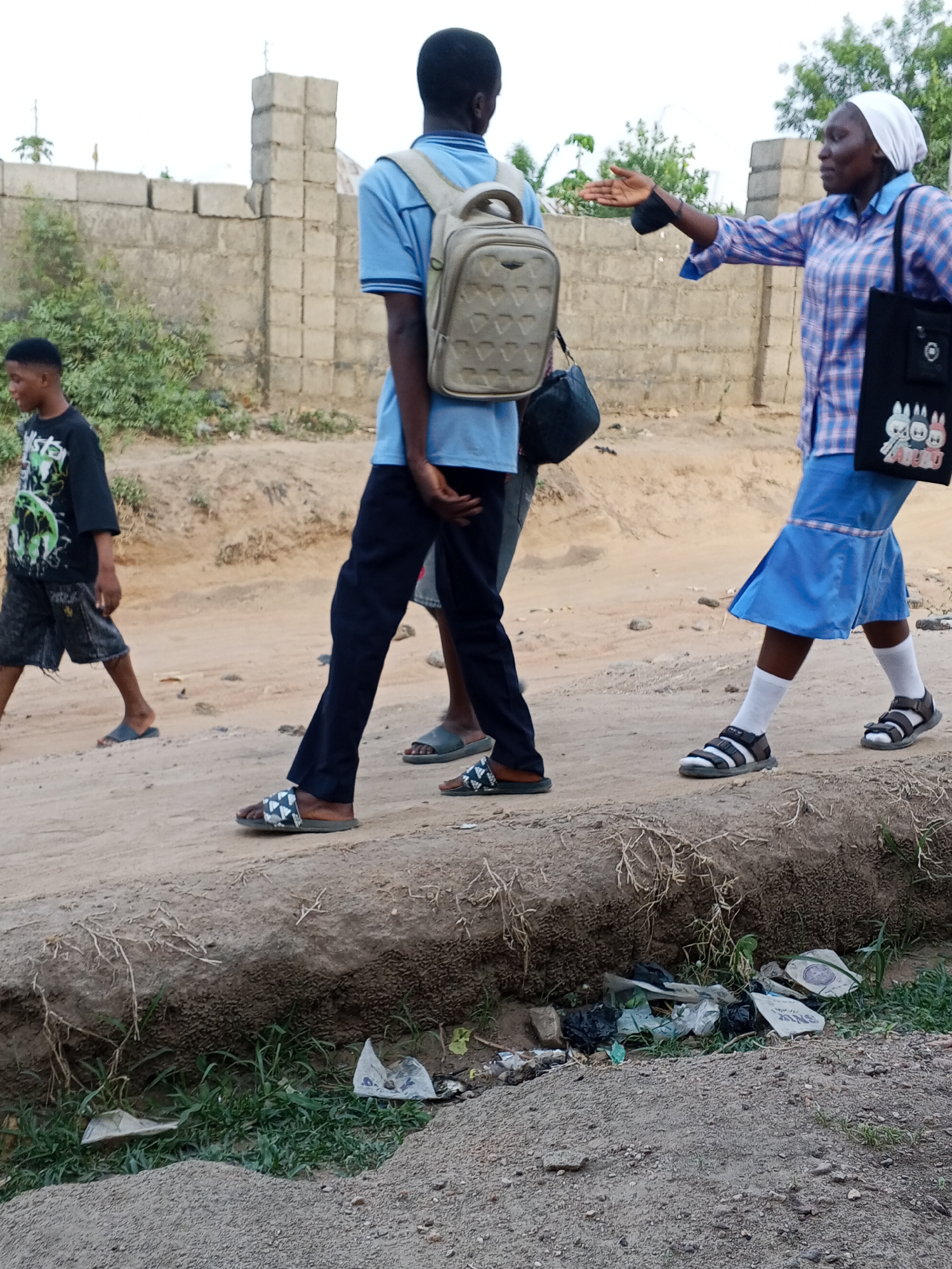 The image depicts a scene in Adonkolo, Nigeria. Three individuals are walking on a dirt path alongside a shallow ditch filled with trash. One person wears a blue polo shirt, dark pants, and sandals, carrying a backpack. A second person follows, and another is walking in the same direction. A woman wearing a checkered shirt, blue skirt, white headscarf, white socks, and sandals extends her arm outward. She carries a black tote bag. A young man in a t-shirt and shorts walks in the opposite direction. A concrete block wall is visible in the background. The environment appears to be a residential or semi-urban area.