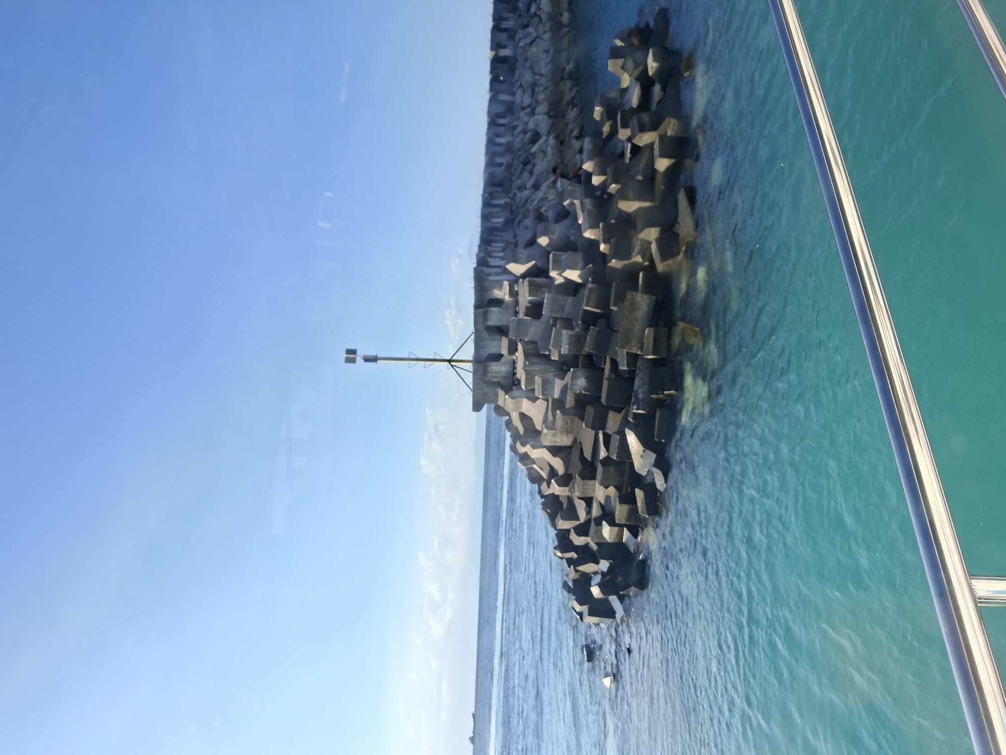 The image depicts a breakwater constructed from large, angular concrete blocks in the ocean off Nusa Penida, Indonesia. The water is a vibrant turquoise, characteristic of the region. In the distance, a hazy landmass is visible on the horizon, possibly other islands or the mainland of Bali. A small cargo ship is present on the ocean. A navigational marker, consisting of a pole with a light fixture and a triangular sign, stands atop the breakwater. The foreground shows a reflective metallic railing, suggesting the photo was taken from a boat or ferry. The sky is clear and blue, indicating fair weather.