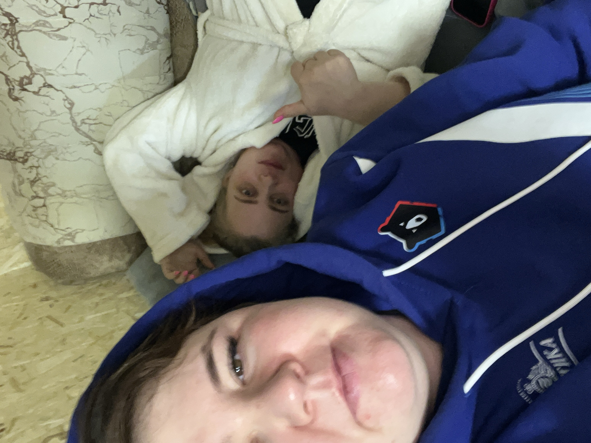 Two individuals are lying down, possibly on a floor or bed. The foreground subject is closer to the camera, wearing a blue zip-up hoodie with white accents and an emblem featuring a bear-like design and Cyrillic text, suggesting a connection to Chaikovsky, Russia. The background subject is wearing a white bathrobe. The environment is illuminated by artificial light. A couch or cushion is visible in the background.