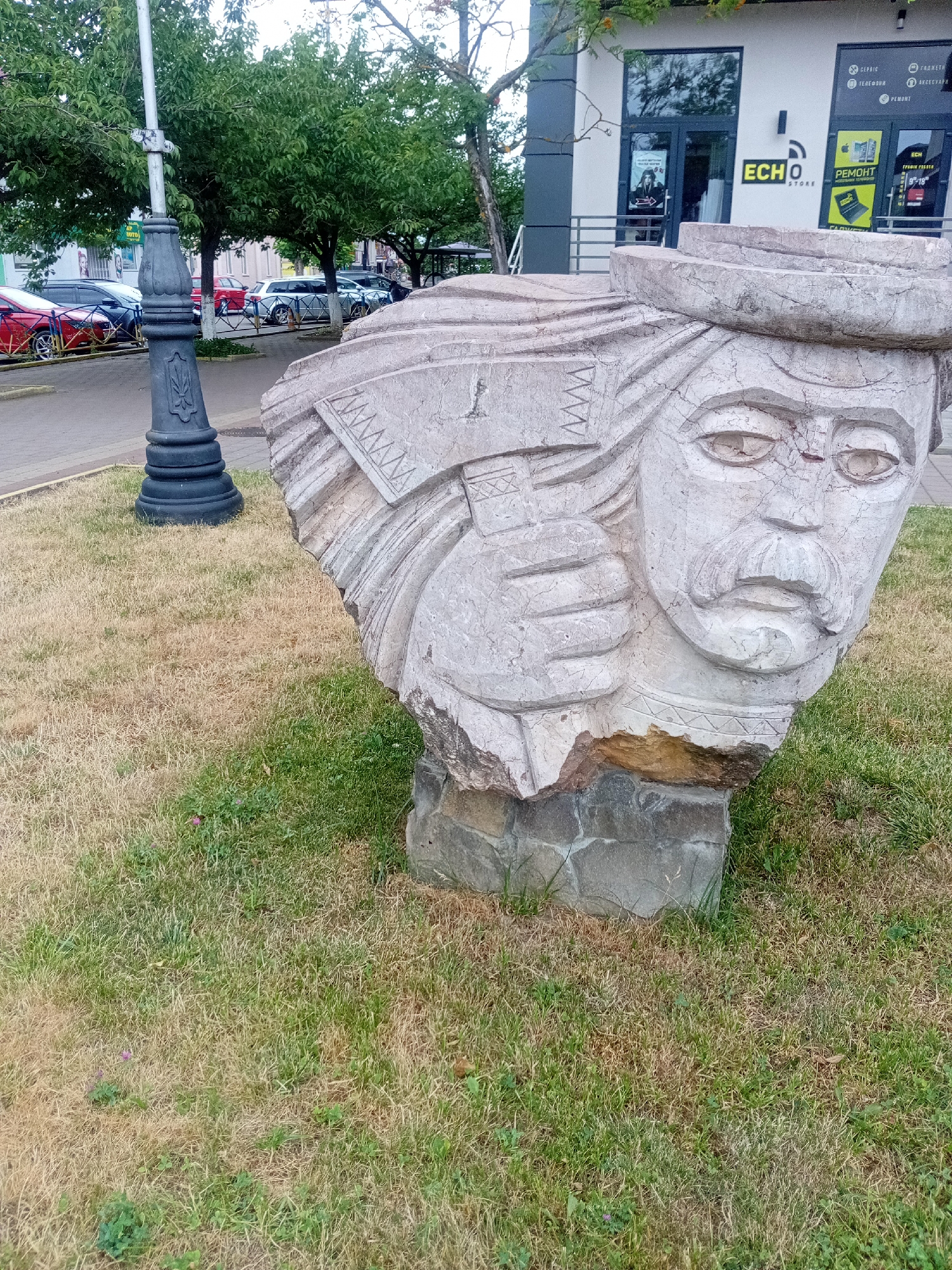 A large, weathered stone sculpture of a man's head with a mustache and a hat is situated on a grassy area in front of a modern building with glass windows and signage. The sculpture has cracked surfaces and is supported by a base of rough stones. To the left of the sculpture, a black decorative lamppost stands on the pavement. In the background, several cars are parked along a street bordered by trees and a metal fence. The building in the background features the logos "ECHO STORE" and advertisements for "РЕМОНТ" (Repair) and "ТЕЛЕФОНИ" (Phones). The grassy area shows patches of dry, brown grass interspersed with green. The overall scene suggests an outdoor public space within a city.
