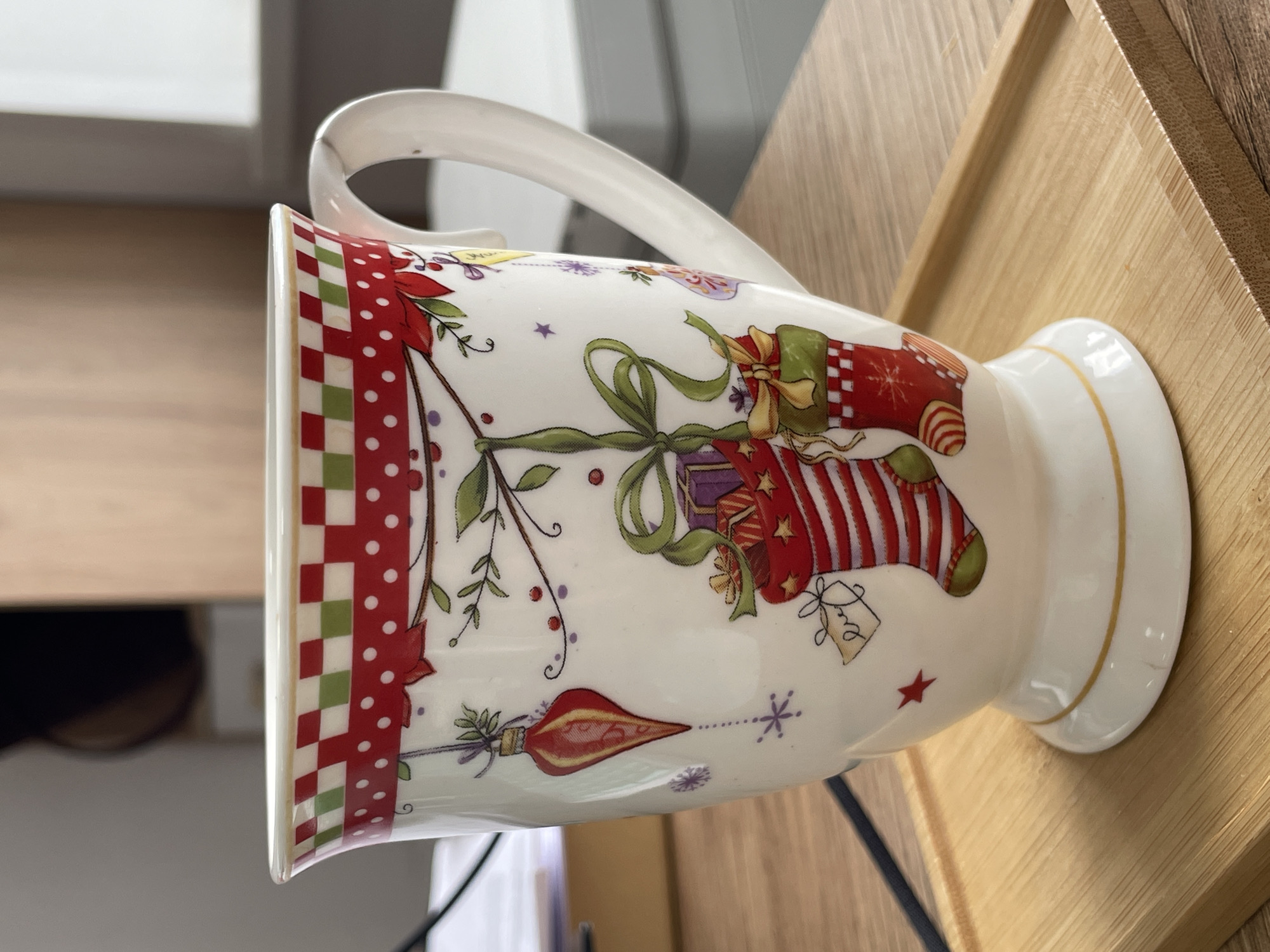 The image displays a white ceramic mug, richly decorated with festive, likely Christmas-themed, illustrations. The mug is positioned on its side on a light-colored wooden surface, which could be a table or a tray. The background is blurred, showing indistinct light-colored shapes, possibly interior walls or furniture.
The mug features a decorative band around its rim, composed of alternating red and green checkered squares alongside red sections with white polka dots. The main body of the mug is adorned with various holiday elements: large red stockings filled with gifts, wrapped present boxes tied with green ribbons and bows, sprigs of green foliage with red berries, and small purple snowflakes or stars. One stocking has green toes and a heel, while another exhibits red and white stripes. A prominent red, teardrop-shaped ornament is also part of the design. The mug has a white handle and a flared base, which is highlighted with a thin gold rim. The surface of the mug appears smooth and glossy.
No specific activity or event is taking place; the image is a static shot of the mug. The lighting is bright and even, suggesting an indoor setting during the day or under artificial light. There is no visible text on the mug or elsewhere in the image. This media file is associated with Izhevsk, Russia.