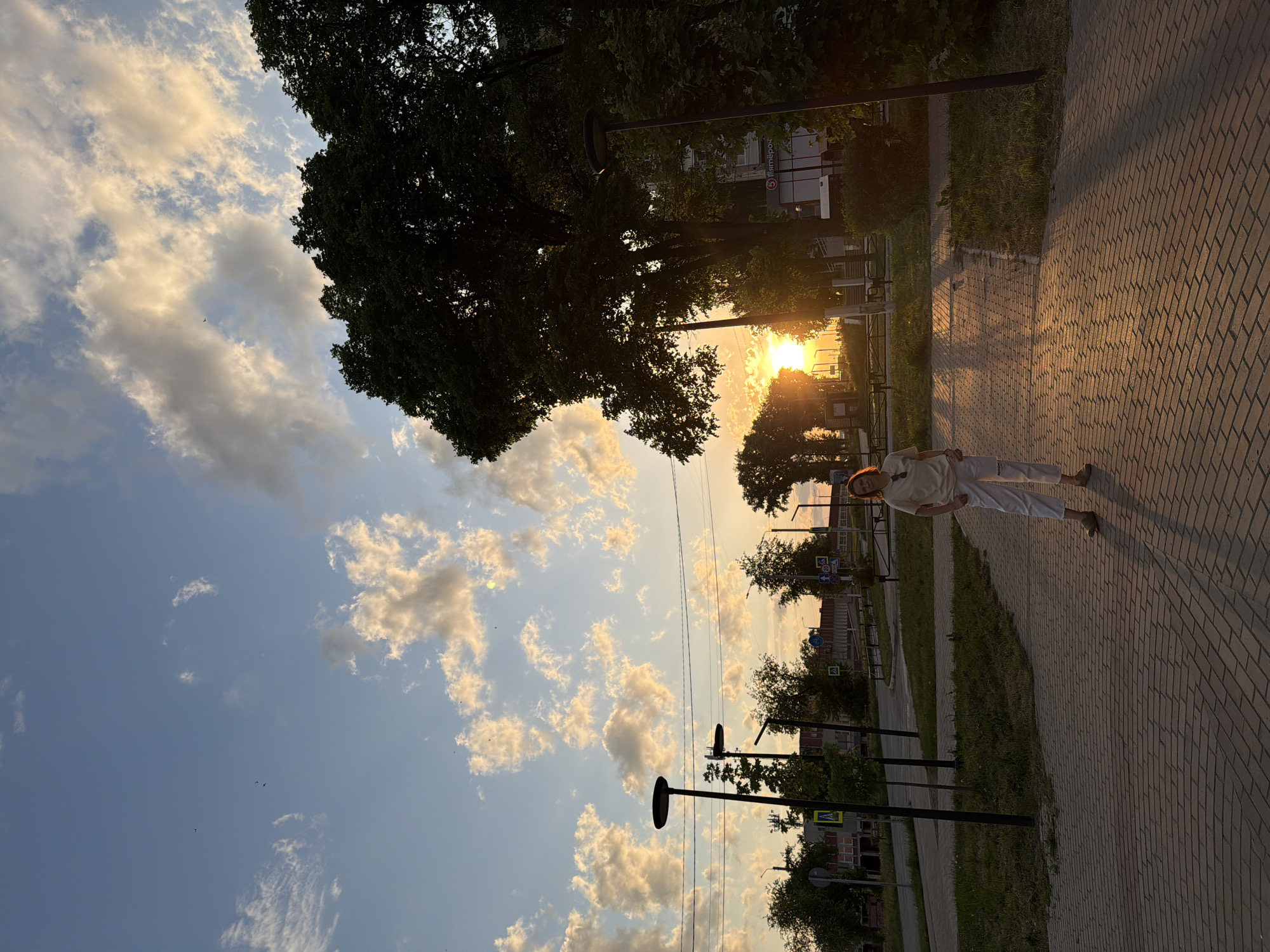 A person is standing on a brick-paved path in the city of Chaikovsky, Russia, during sunset. The path is lined with grass and small trees, and modern lampposts are spaced along the edge of the walkway. Buildings are visible in the background. The sky is a mix of blue and cloudy hues with the sun setting in the center of the frame.