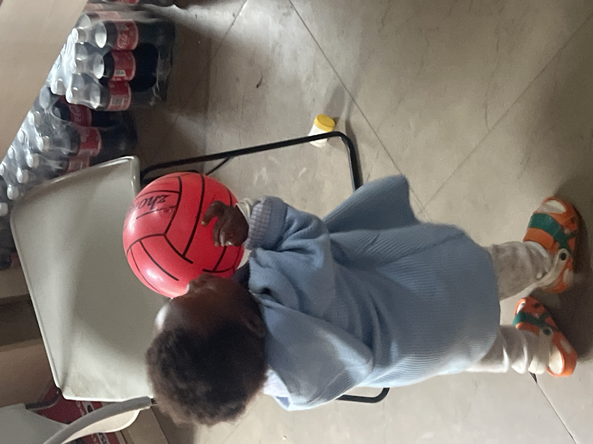 A child wearing orange and green sandals, white pants, and a light blue sweater is lying on a light gray tile floor. The child is holding a red ball with black lines. Next to the child, on a chair, are packs of bottled drinks. A small yellow container is on the floor near the chair. The location is not identifiable.