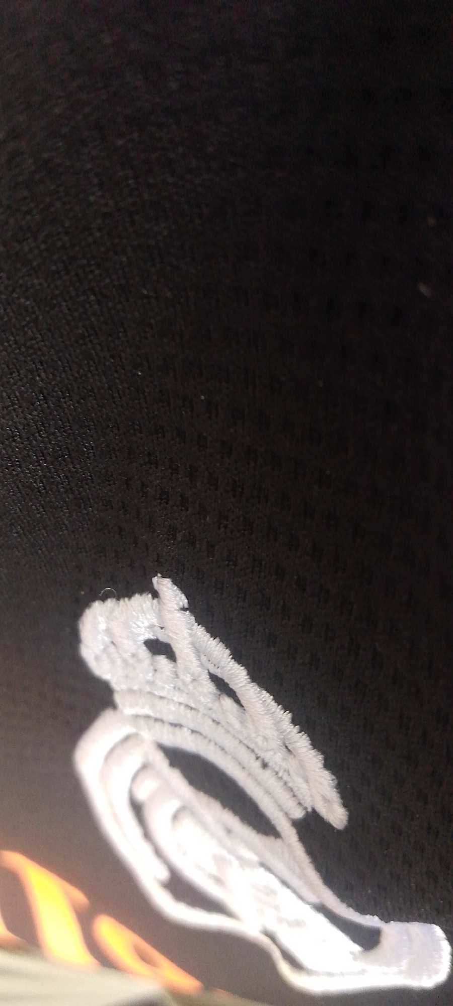 A close-up, angled shot of a white embroidered design on black fabric. The embroidery appears to be a stylized abstract pattern with swirling lines and loops, possibly part of a logo or decorative element. The fabric has a textured surface, visible as small, uniform dots. To the lower left, a blurred area of warm, orange and yellow light suggests an external light source or reflection, though its origin is unclear. The image provides no contextual information about its location in Jalingo, Nigeria.