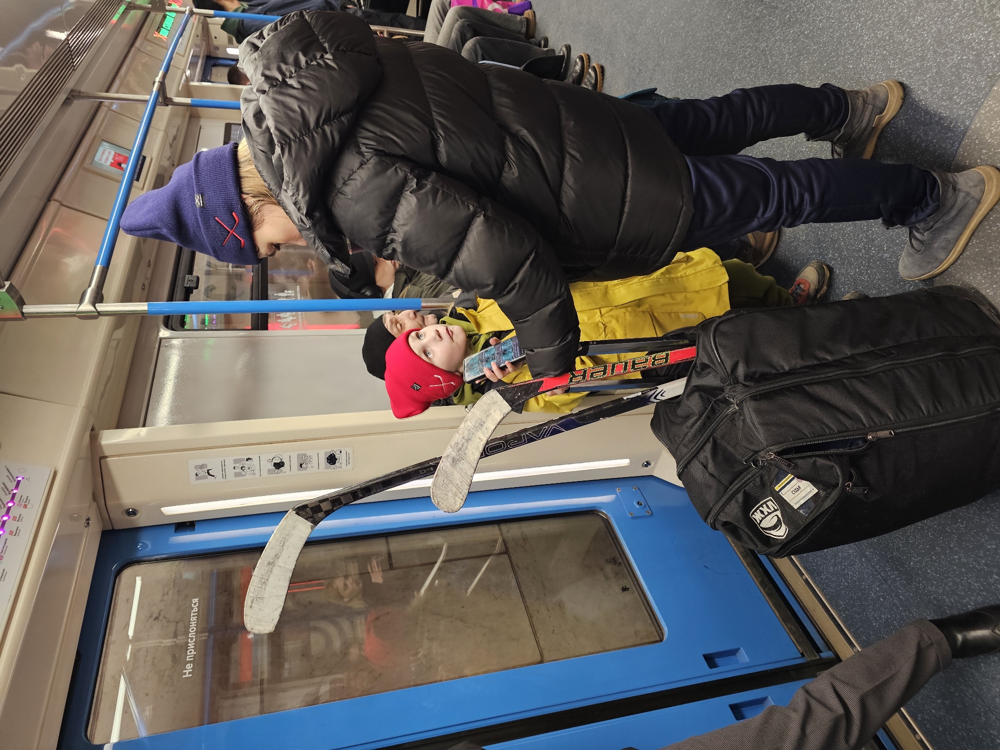 The image shows the interior of a Moscow Metro train in Russia. A person wearing a black puffer jacket, blue pants, and gray boots stands over a child wearing a yellow jacket and a red hat. The child is looking at a smartphone. Next to them are two hockey sticks and a black equipment bag. The train car has blue doors and cream-colored paneling with informational stickers. The floor is gray. Other passengers are seated in the background.