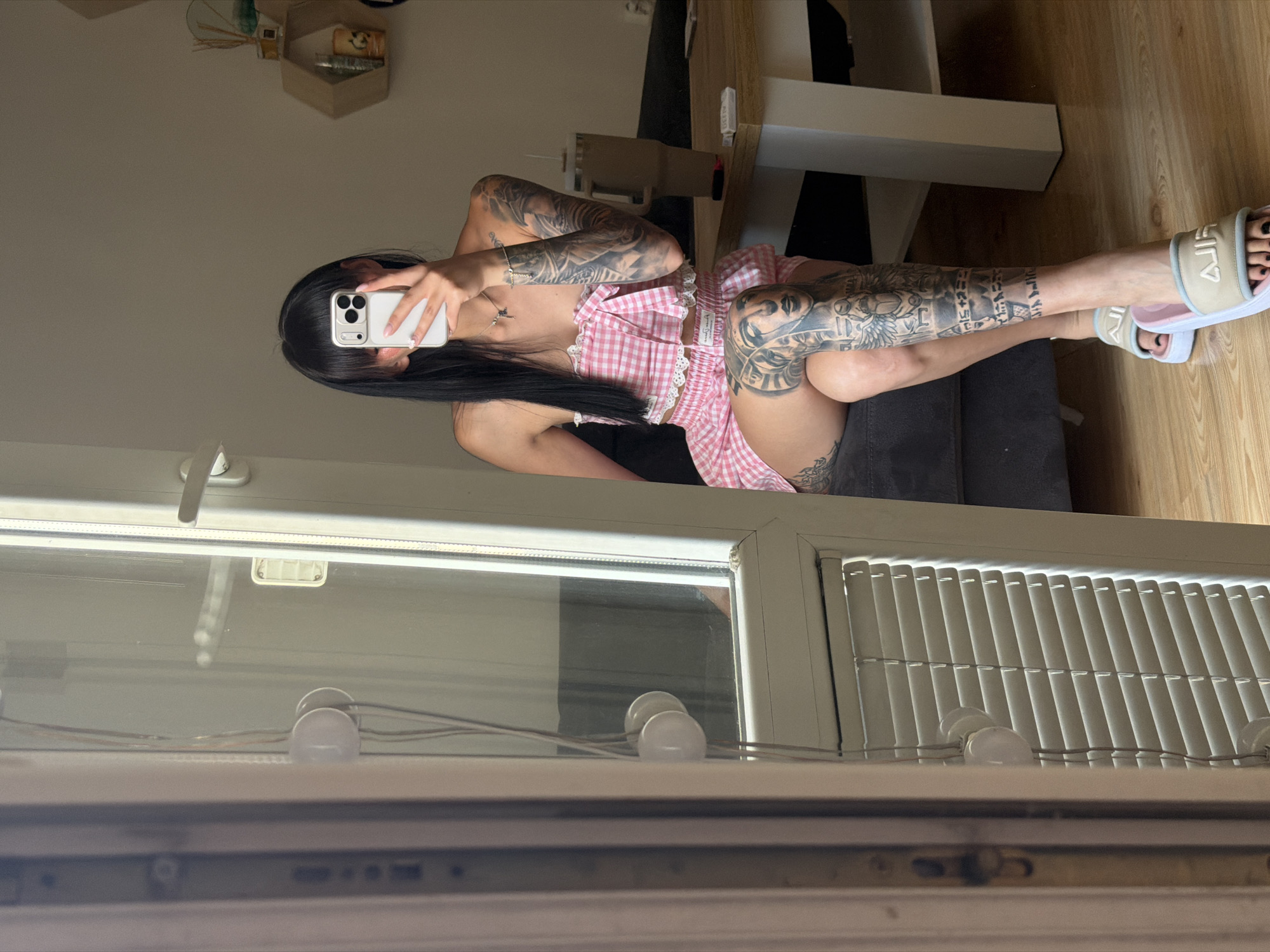 A young woman with long black hair and bangs is sitting indoors, taking a mirror selfie with her phone. She is wearing a pink gingham tube top and matching shorts. Her arms and legs are covered in extensive black and grey tattoos. She is wearing light-colored slides with a pink trim. The background shows a reflective surface that appears to be a door or window, with blinds on the lower portion. A wooden table with a tall cup and a small object is visible in the right side of the frame. The environment suggests an indoor setting, possibly a residential room, with a neutral-colored wall and wooden flooring. The lighting appears to be natural, coming from the right side. There are no specific landmarks or indications of the city of Plovdiv, Bulgaria, in the image.