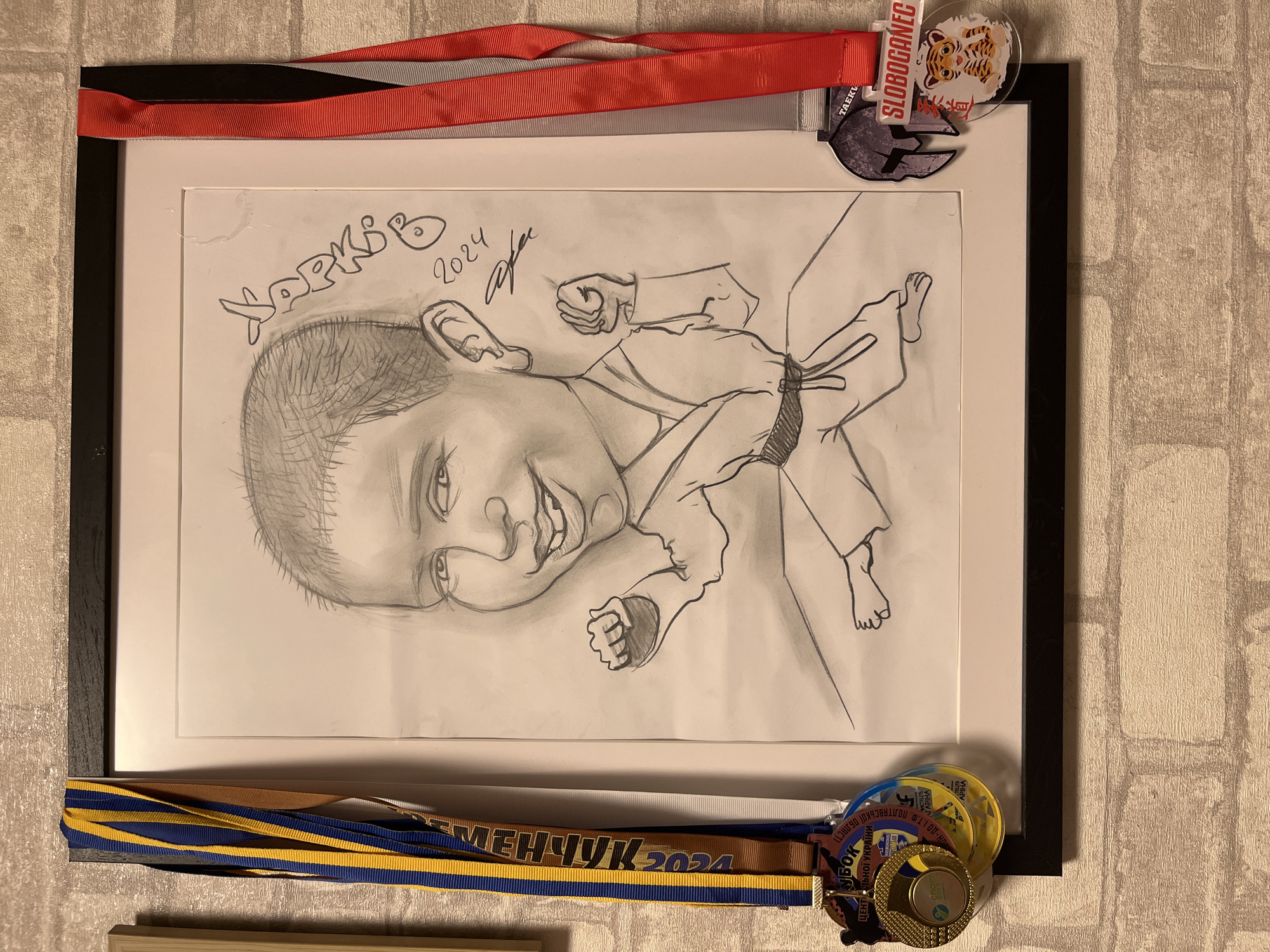 The image shows a framed caricature of a young boy in a karate gi, drawn in pencil. The boy is depicted in a dynamic pose, with his fists raised. The drawing is signed "XAPKO" and dated "2024" with a stylized signature below. The frame is black and is hanging on a light-colored brick wall.
Several medals are attached to the frame. On the left side, there are three medals with blue, yellow, and brown ribbons. One of these medals has Cyrillic text that appears to mention "UKRAINE" and "2024". On the right side, there are two medals with red and gray ribbons. One of these medals has the text "TAEKWONDO" and "SLOBOGANEC" printed on it, along with a graphic of a tiger.
The location of this scene is not explicitly mentioned in the image itself, and therefore cannot be determined.