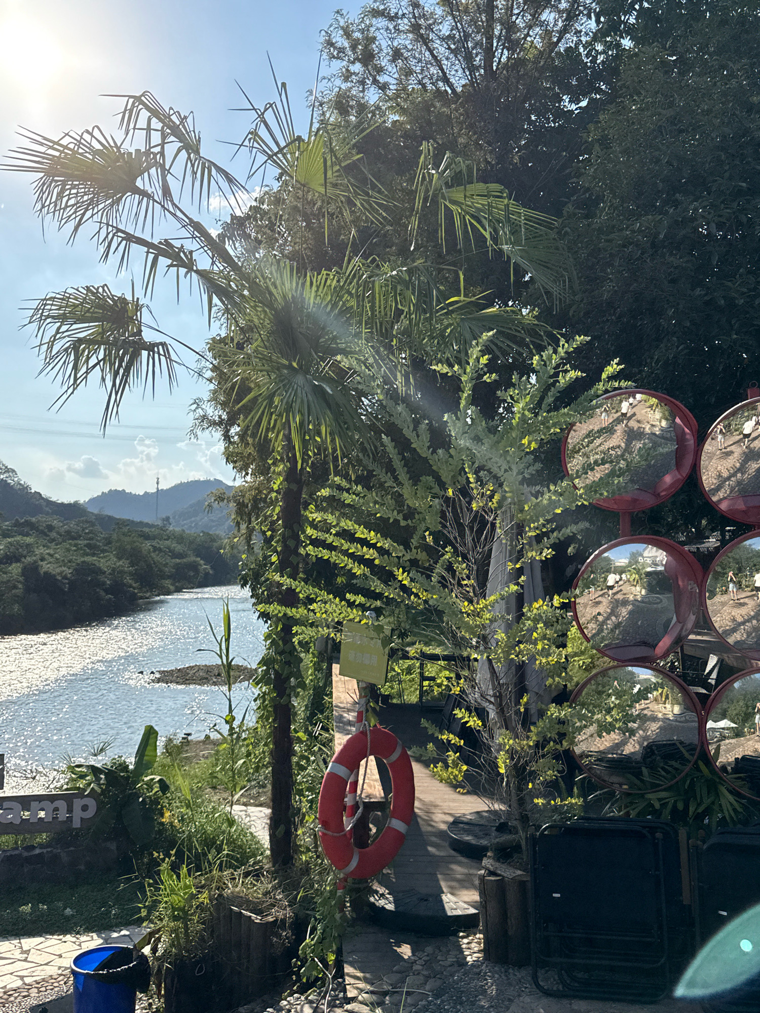 The image depicts a serene riverside scene with lush greenery and a bright, sunny sky. In the foreground, a red life preserver hangs on a wooden post adorned with climbing vines. To the left, a blue trash can sits on a paved walkway. A wooden sign with the word "camp" is partially visible, indicating a camping area. Several round, convex mirrors with red frames are mounted on a pole, reflecting the surrounding environment and a group of people walking. Beyond the immediate foreground, a tranquil river flows, its surface shimmering in the sunlight. The riverbanks are lined with dense vegetation, and in the distance, rolling hills covered in trees recede into the horizon under a partly cloudy sky. The overall impression is one of a peaceful outdoor recreational space. This scene is located at the Yiwu River Scenic Area in Yiwu City, China, a popular destination for leisure and outdoor activities.