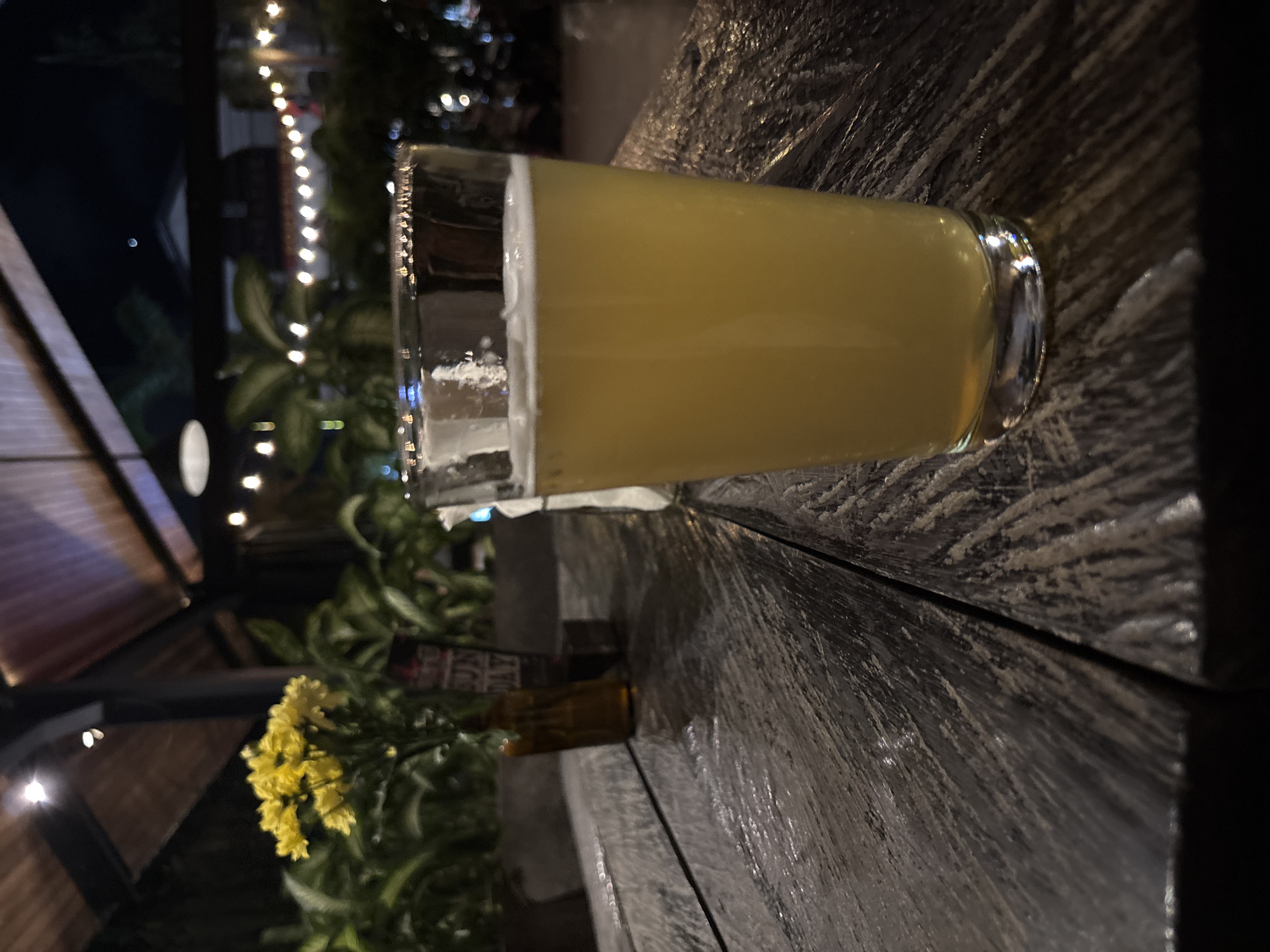 A glass of beer with approximately one inch of foam rests on a dark, rough-hewn wooden table. In the background, out-of-focus lights suggest an outdoor or semi-outdoor setting. A small vase of yellow flowers is visible to the left, also slightly out of focus. The lighting indicates it is evening or nighttime. The location is North Kuta, Indonesia.