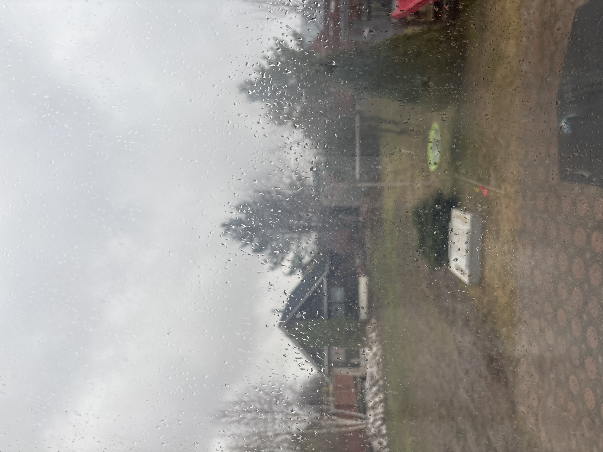 The image depicts a view through a rain-streaked window. Outside, a house with a dark roof is visible amidst trees and foliage, likely in a residential area. To the right, there appears to be a paved area with a patterned surface. A green object, possibly a kayak, is also visible. The overall scene suggests a rainy day. Location cannot be verified.