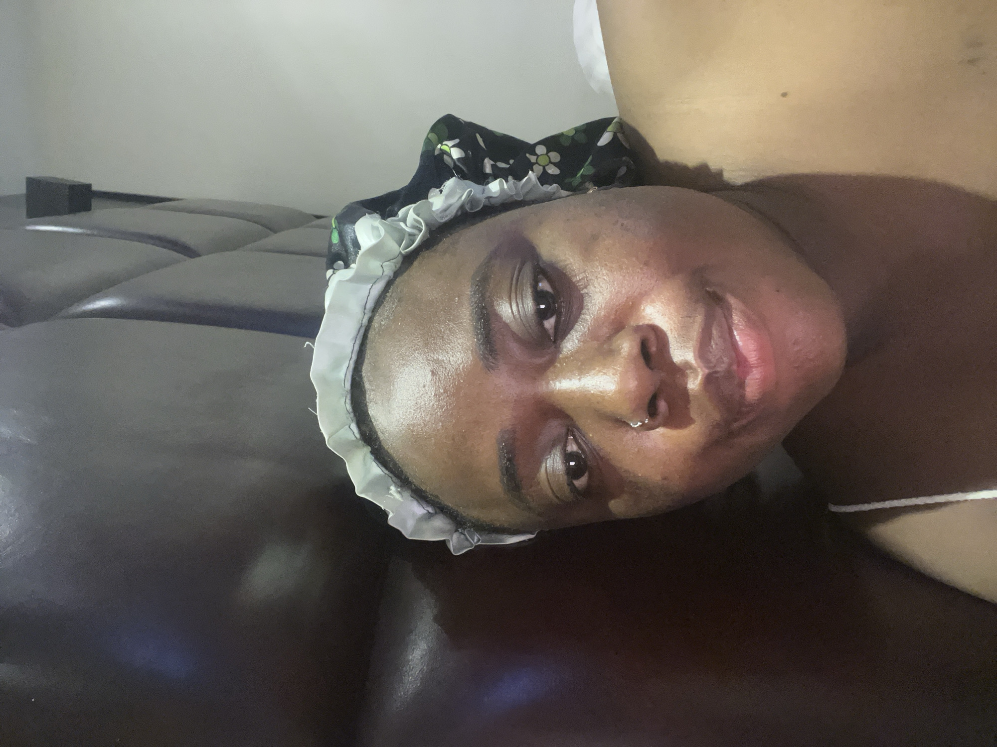 A close-up shot of a person's face. The person wears a black floral bonnet with white ruffles. They have a nose ring. The person is lying on a brown, tufted leather surface. A dark rectangular object sits behind the person's head. The location is not discernible from the image, and I cannot determine if it is in Abuja, Nigeria.