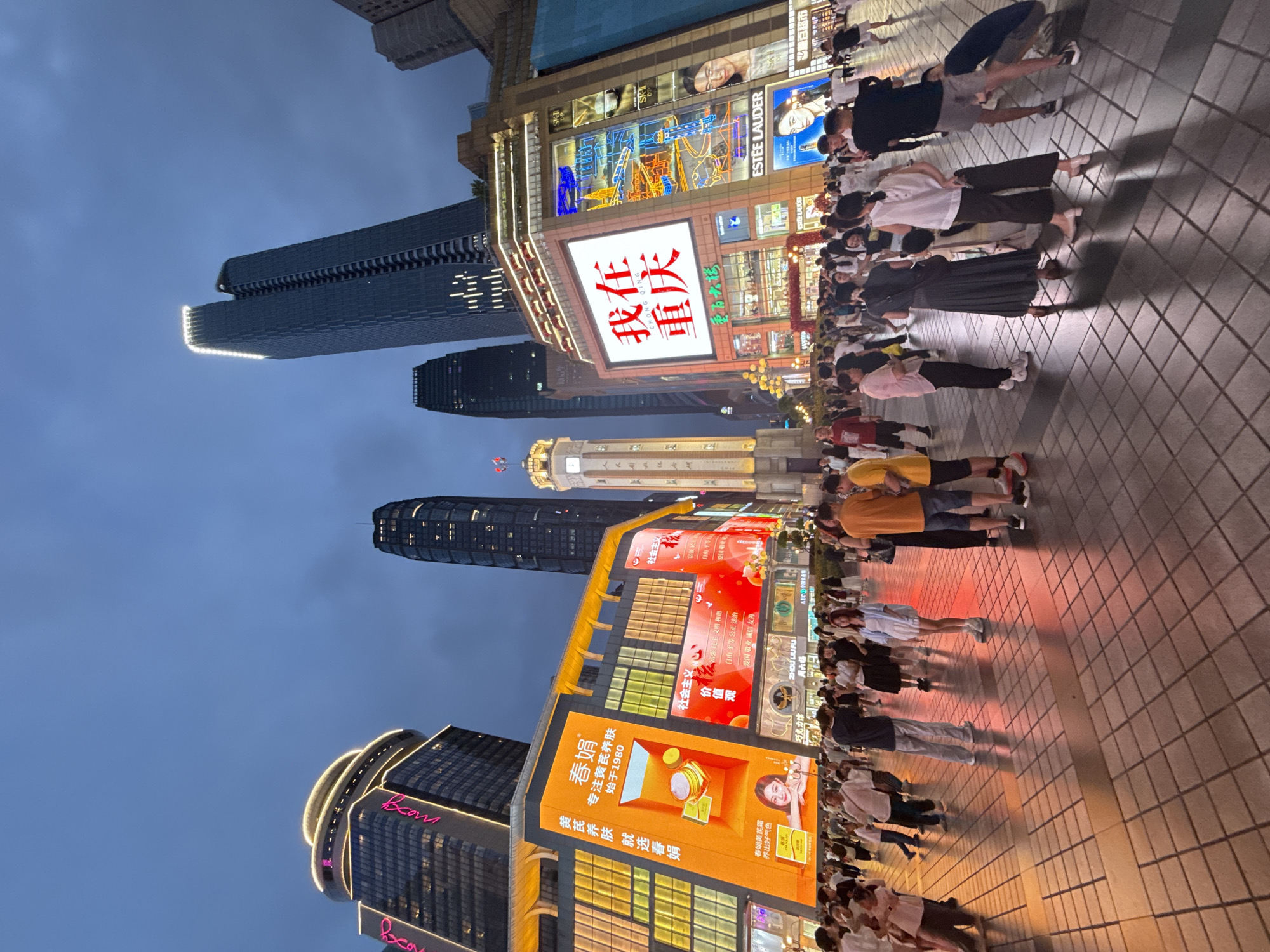 In Yuzhong District, China, this nighttime urban scene depicts a bustling pedestrian area at dusk. Towering skyscrapers, illuminated by strings of lights and large digital advertisements, dominate the skyline. One building features a prominent digital billboard displaying red Chinese characters that translate to "I am in Chongqing." Another billboard advertises a skincare brand.
A clock tower stands prominently in the mid-ground, with people gathered around its base. In the foreground, a vast tiled plaza is filled with a diverse crowd of people, dressed in casual summer attire, engaged in various activities. Some are walking, others are standing in groups, and a few are resting. The ground is tiled with a grid pattern, reflecting the ambient light from the surrounding buildings and billboards. The overall atmosphere is lively and vibrant, indicative of a popular urban gathering spot.