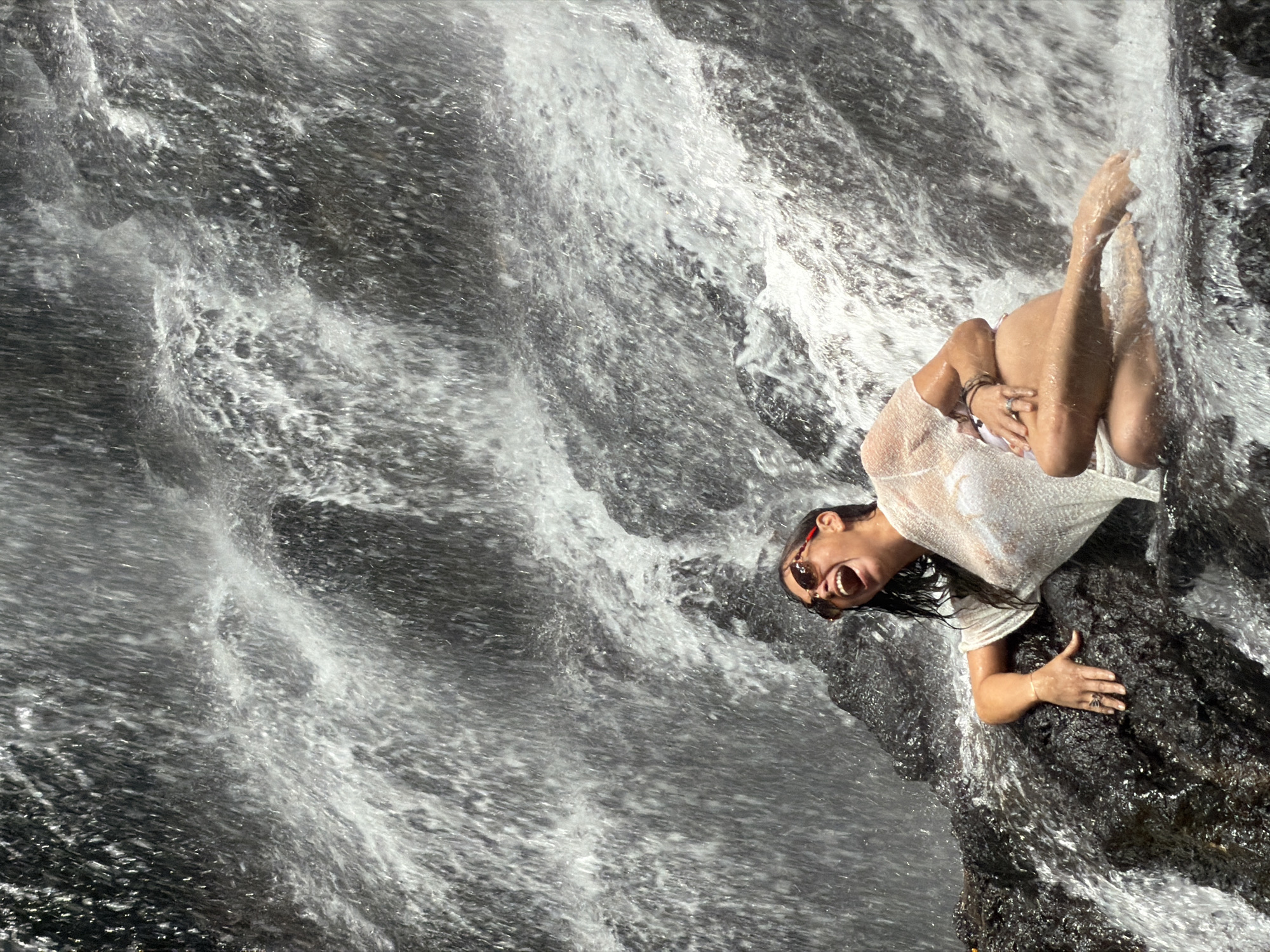 A woman with wet hair and wearing sunglasses and a sheer white top over a bikini sits on dark, wet rocks in front of a cascading waterfall. She is laughing with her mouth open and her head tilted back, with her left arm resting on the rock and her right arm bent across her chest. Her legs are bent and submerged in the water. The environment appears to be a natural setting with a significant amount of flowing water and dark, textured rocks. The scene is captured in a vertical, close-up shot, focusing on the woman and the immediate surroundings of the waterfall. The specific location in Gianyar, Indonesia, is not discernible from the visual information alone.