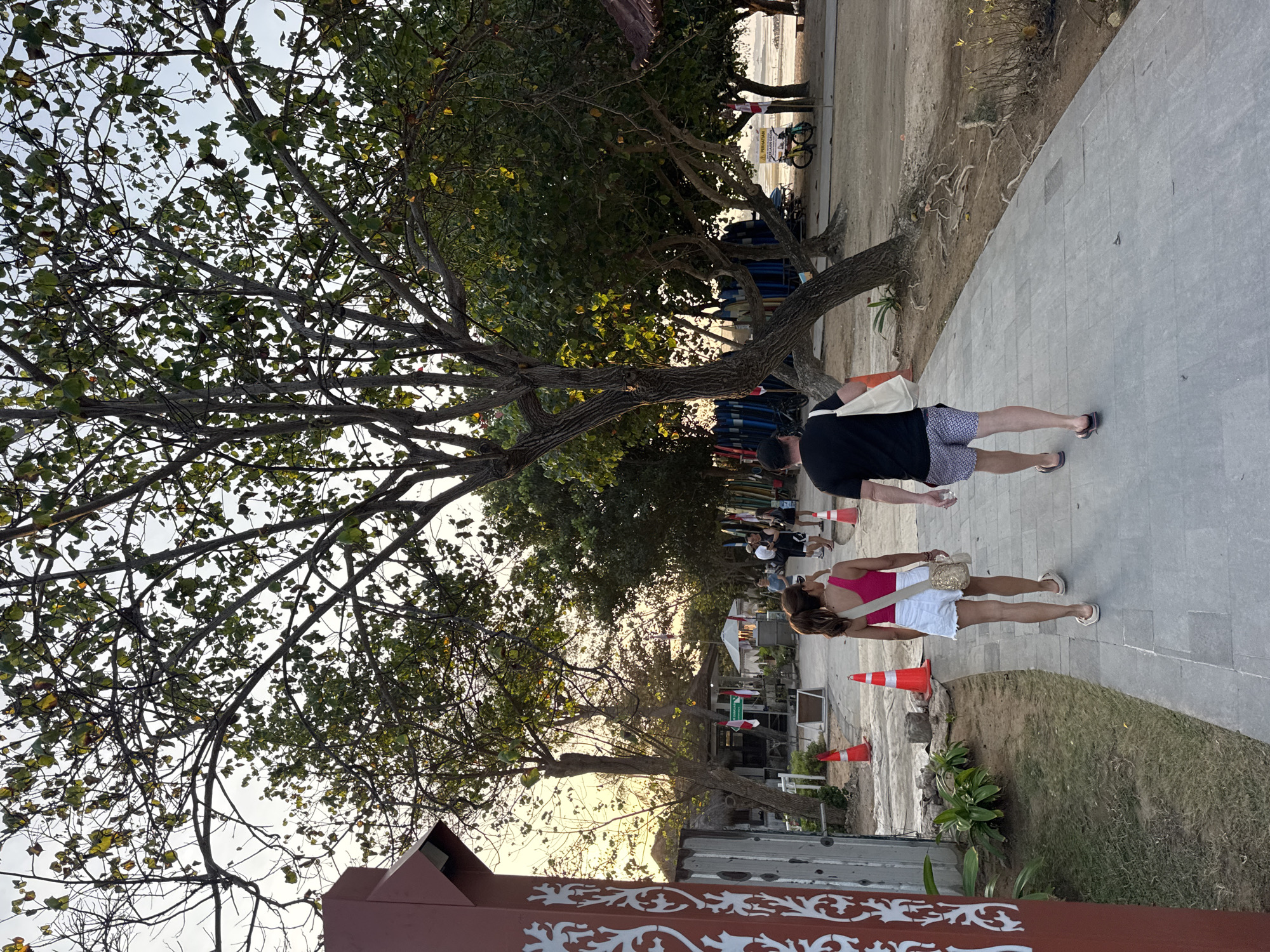 In Kuta Selatan, Indonesia, a paved pathway curves through a sandy area dotted with trees and vegetation. A couple walks away from the camera on the pathway. The woman, in a bright pink tank top and white shorts, carries a tan purse. The man, wearing a black t-shirt and patterned shorts, carries a white tote bag. In the background, several surfboards are propped upright near a small building. Orange traffic cones are placed along the side of the pathway, and a bicycle is visible further down the path. The sky is overcast with the warm glow of the setting sun visible through the trees.