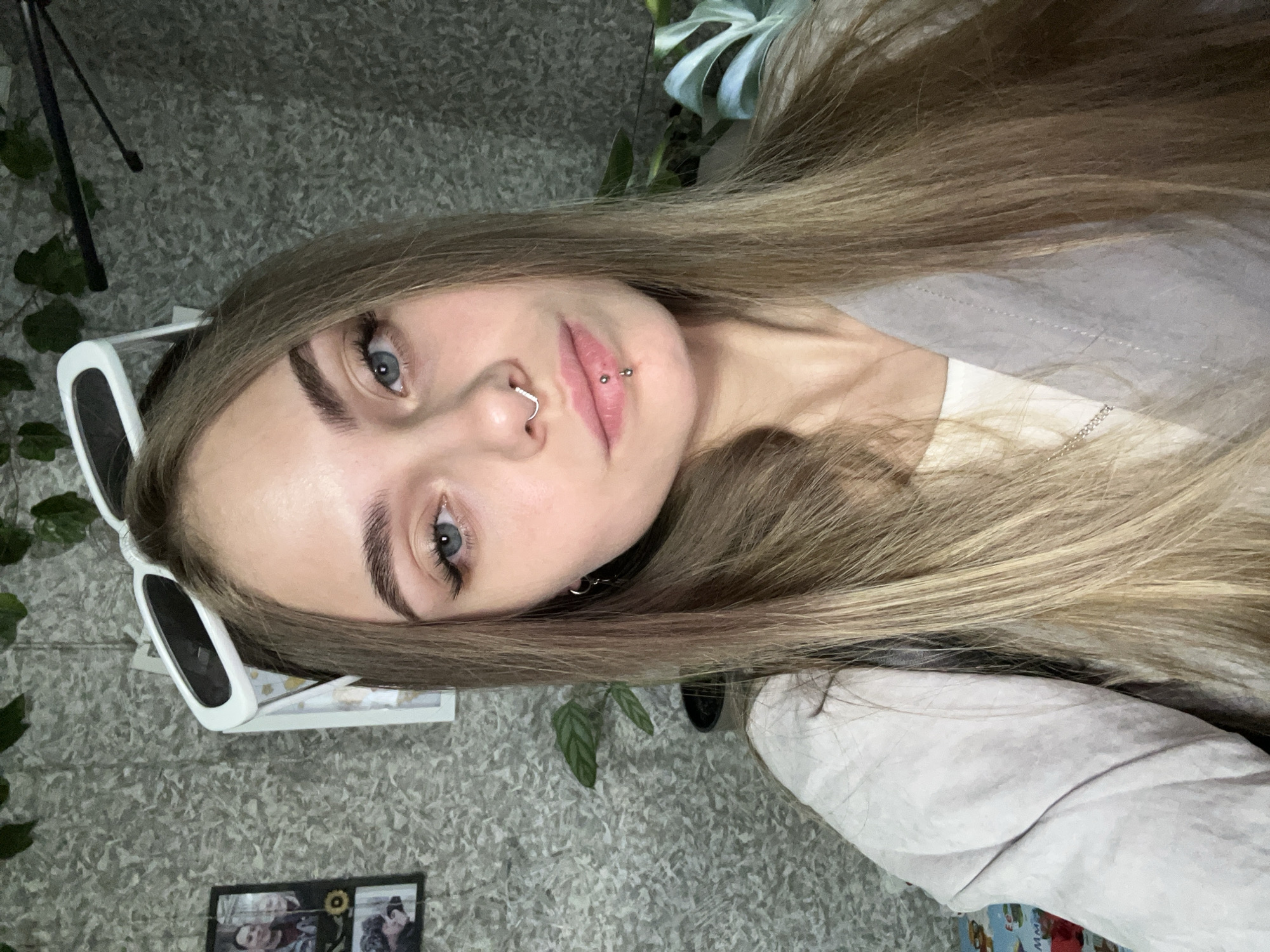 A young woman with blonde hair and blue eyes is pictured indoors. She has piercings in her nose and lip. She is wearing a light-colored shirt. White sunglasses are perched on her head. The background includes a patterned gray wall, some green plants, and a tripod. The photo seems to be taken indoors. The image doesn't include any identifying information to confirm the location is in the city of Фоки, Russia.