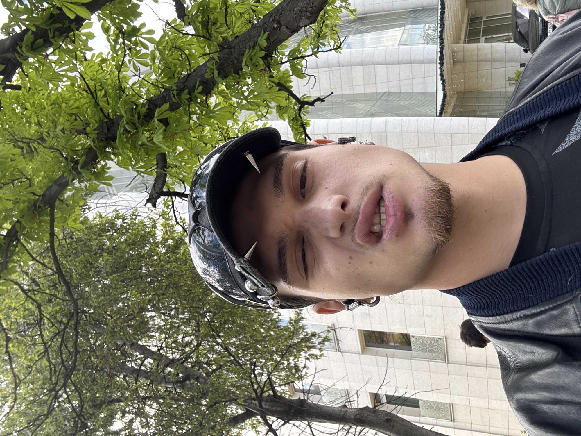 This vertical-format image, rotated counter-clockwise, captures a low-angle selfie of a young person outdoors in Sofia, Bulgaria. The subject, likely male with short dark hair and light stubble on their chin and jaw, dominates the foreground. They wear a distinctive black visor or hat embellished with prominent silver spikes and metal studs. Their left ear is adorned with multiple metallic piercings, including various hoops and studs. The person is dressed in a black t-shirt and a dark, possibly leather, jacket featuring a ribbed dark blue collar. Their expression is stylized, with their mouth slightly open, revealing teeth, and lips subtly pursed, while one eye appears slightly squinted.
The background is a blend of nature and urban architecture. Lush, vibrant green leaves of large trees are visible against a bright sky, suggesting a spring or early summer setting. Adjacent to the trees are modern buildings with clean, light-colored facades and numerous rectangular windows, some reflecting the bright sky or displaying subtle patterns. The clear, bright lighting indicates it is daytime. The image presents an edgy, personal portrait against a backdrop of city life. No text is visible in the image.