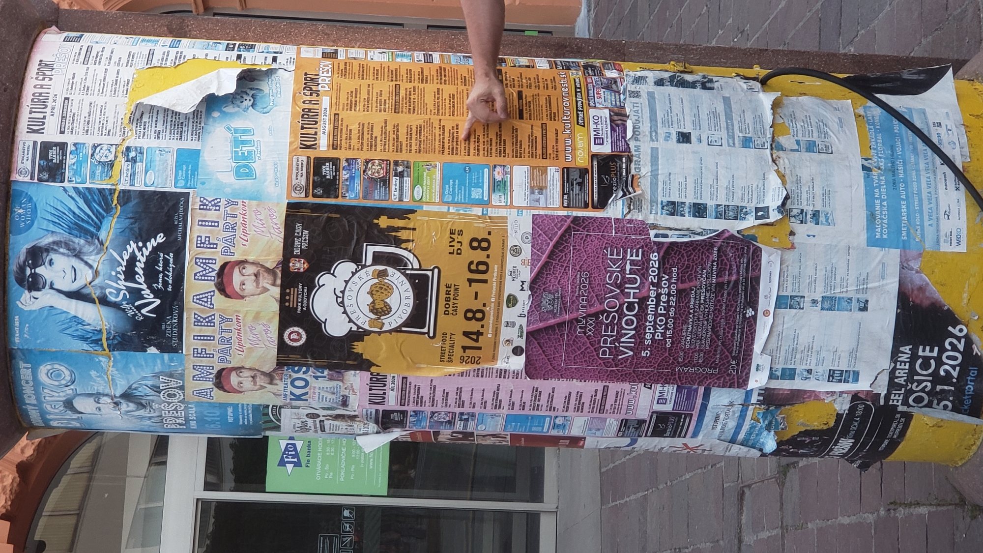 This image shows a lamppost covered in posters in the city of Prešov, Slovakia. The posters advertise various events, including concerts, parties, and cultural events, with dates ranging from 2026. Some of the posters feature prominent Slovak personalities such as Shirley Valentine and Miro Jaroš. The lamppost is situated outdoors, with a building visible in the background and a paved walkway in the foreground. The posters are layered on top of each other, with some torn and weathered, indicating the passage of time and exposure to the elements. A hand is visible reaching out to one of the posters, possibly pointing at specific information. The overall scene suggests a public space where local events and advertisements are displayed to the community.