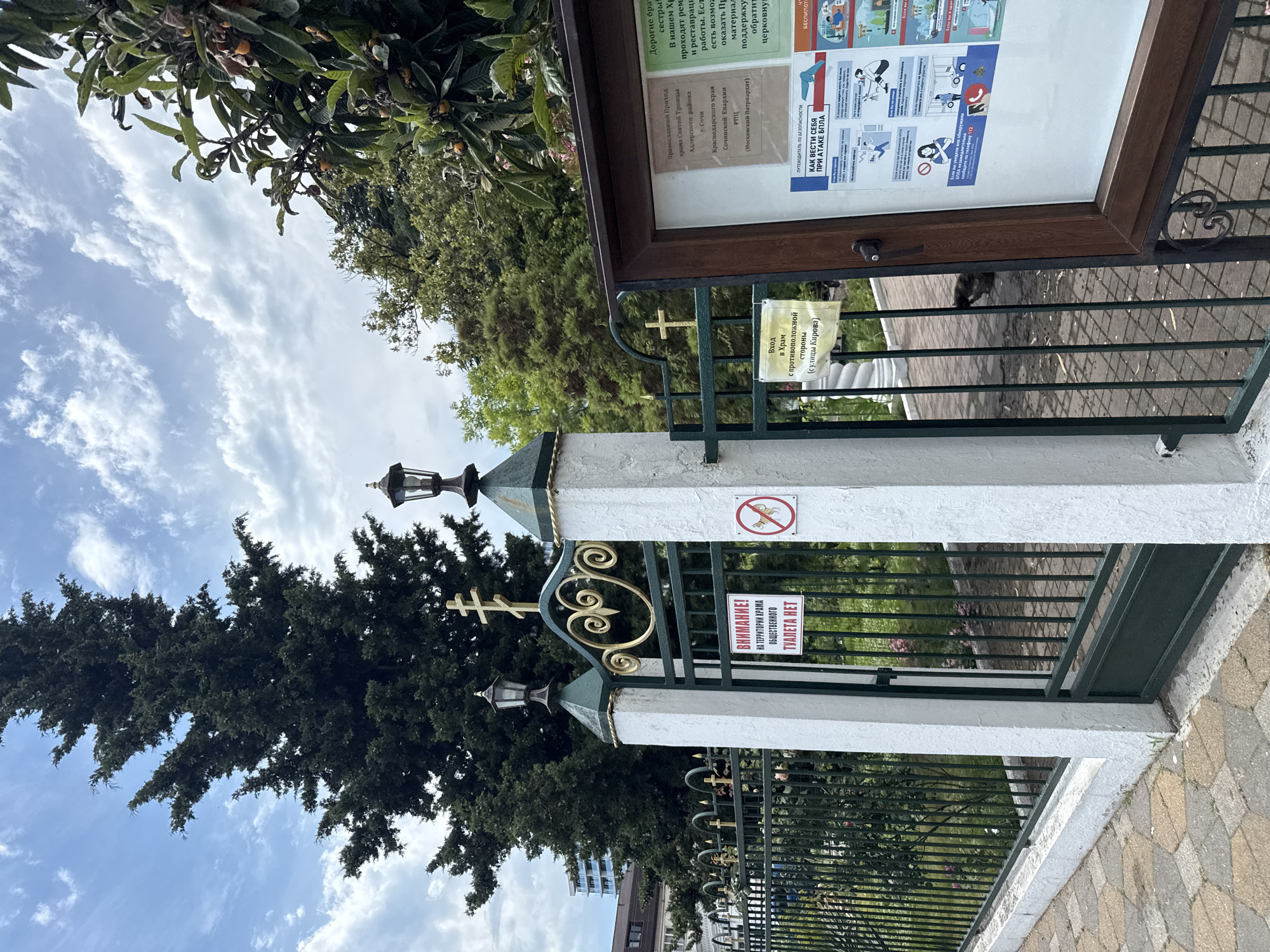The image shows a gate with a sign in Russian reading "ATTENTION! THERE IS NO PUBLIC TOILET ON THE TERRITORY OF THE CHURCH." A red circle with a diagonal line across it and a dog inside indicates that dogs are not allowed. The gate is green with ornate gold detailing and topped with crosses. The gate is flanked by white pillars with lantern fixtures. Behind the gate, a fence encloses a green area with trees and some pink flowers. In the background, a tall evergreen tree and lush foliage are visible, along with a modern building. A bulletin board with various notices, some in Russian, is to the right of the gate. One notice is titled "HOW TO BEHAVE DURING A BILLET ATTACK." The pavement in front of the gate is made of hexagonal stones. The overall scene appears to be the entrance to a church or religious complex in Sochi, Russia.