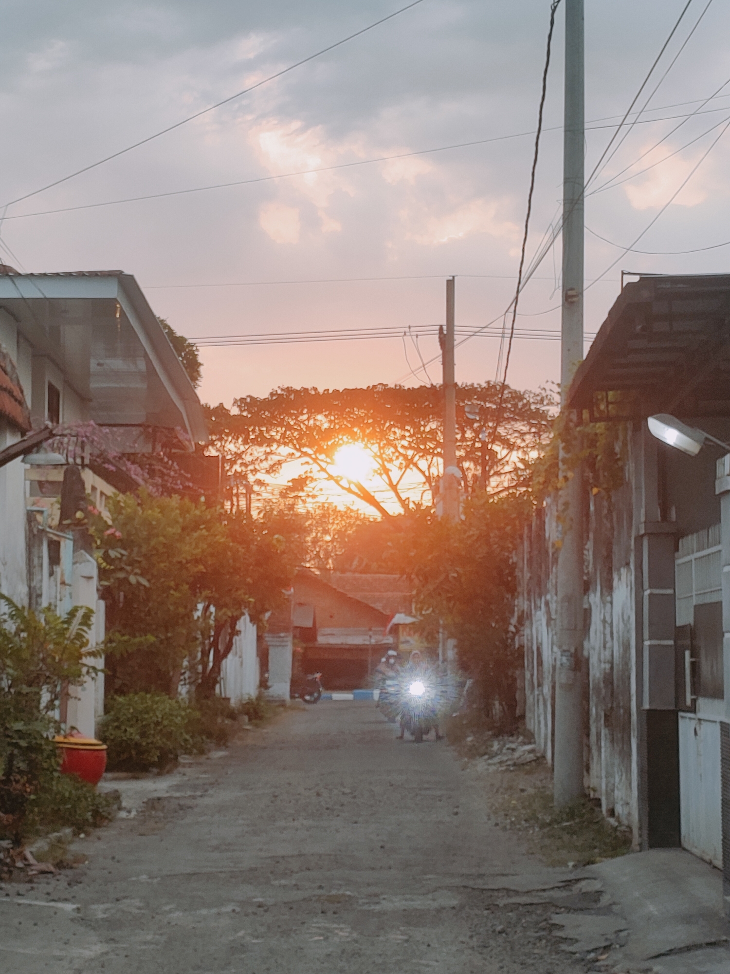 The image depicts a street scene at sunset in Sooko, Indonesia. The sun is low in the sky, casting a warm, orange glow through the trees and clouds. A paved road runs through the center of the frame, lined with houses and vegetation on either side. In the distance, a person is riding a motorcycle, their headlight shining brightly. Overhead, power lines crisscross the sky, supported by utility poles. The overall impression is one of a quiet, residential area as dusk settles.