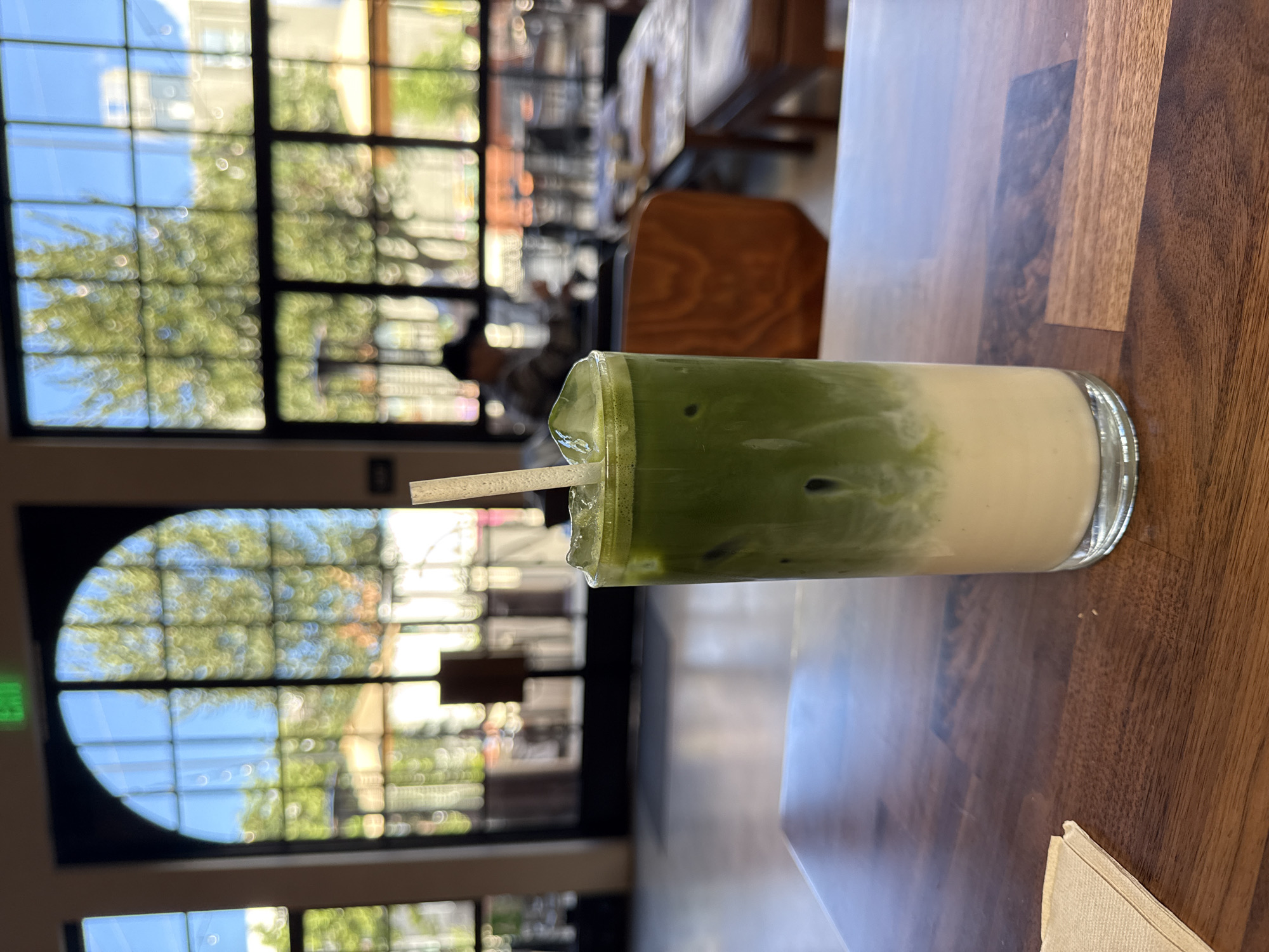 The image shows a partially consumed matcha latte drink in a clear glass, with a straw, set on a wooden table. The drink appears to be in a cafe or restaurant setting. Through the windows, there is a blurred view of trees and buildings. The environment suggests a daytime scene, likely sunny, with people visible in the background. It is impossible to confirm if the location is Los Angeles without any information.