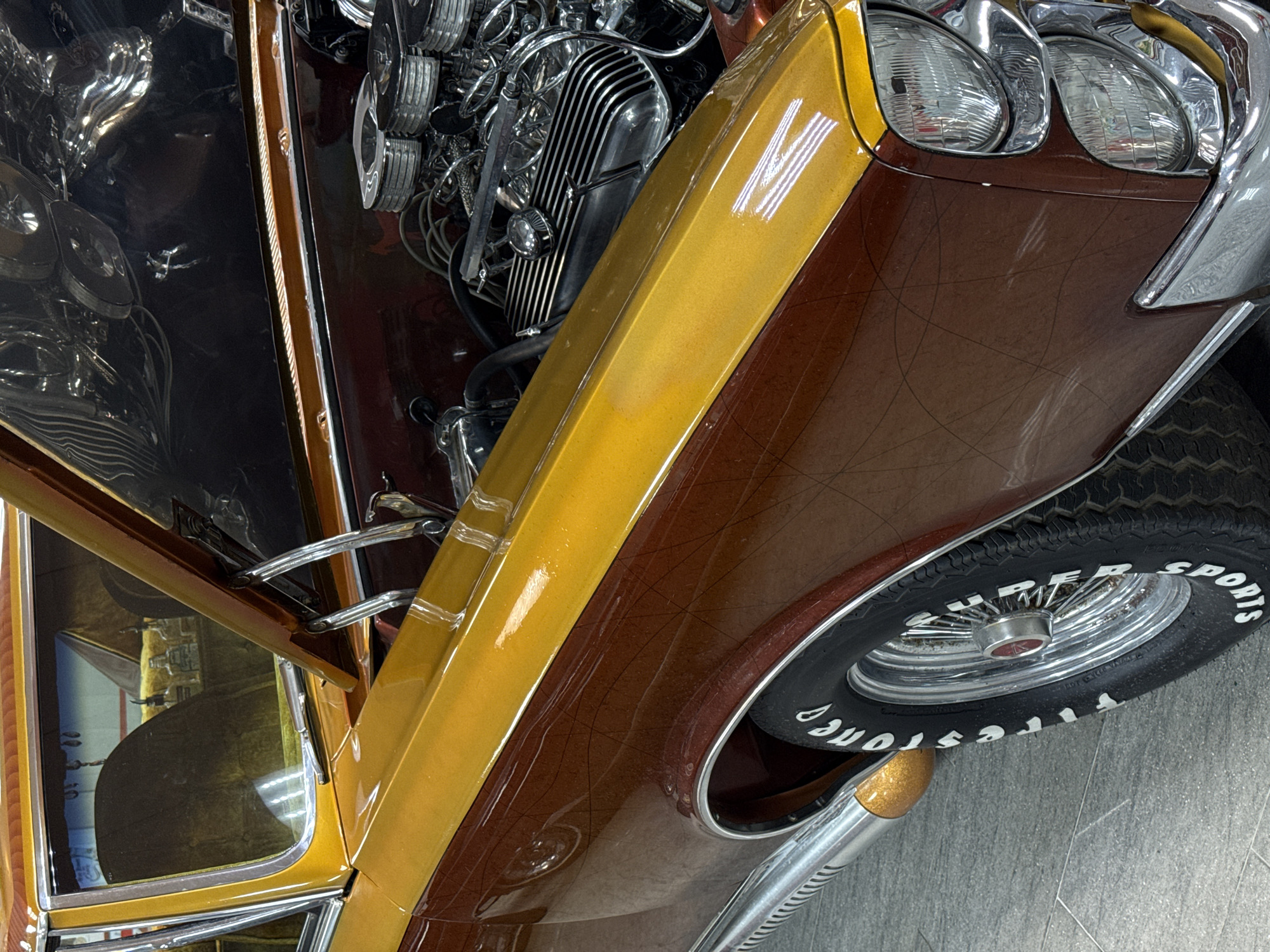 An eye-level shot captures a highly customized or classic car, presented indoors in what appears to be a showroom, garage, or exhibition space in Sochi, Russia. The vehicle boasts a striking two-tone paint job, featuring a vibrant, metallic golden yellow stripe running along its side, complementing a rich, shimmering metallic brown body. The brown body panels are adorned with a subtle, intricate pattern of intersecting lines, possibly etched or pinstriped.
The car's hood is partially open, revealing a meticulously detailed engine bay. The engine, likely a V8, is heavily chromed, with polished valve covers, multiple air intakes, and intricate plumbing, characteristic of a show car. The driver's side door is also ajar, providing a glimpse of a custom interior with upholstery in a golden-yellow hue.
The front of the car features dual round headlights encased in chrome bezels. The front wheel is equipped with a black wall tire clearly labeled "FIRESTONE SUPER SPORTS" in white lettering, mounted on a chrome wire spoke rim with a red center cap. A prominent, polished chrome side exhaust pipe is visible beneath the door. The car rests on a clean, light grey tiled floor, which reflects the vehicle's polished surfaces and the artificial overhead lighting. The stationary car, with its open hood and door, suggests it is either on display or undergoing preparation.