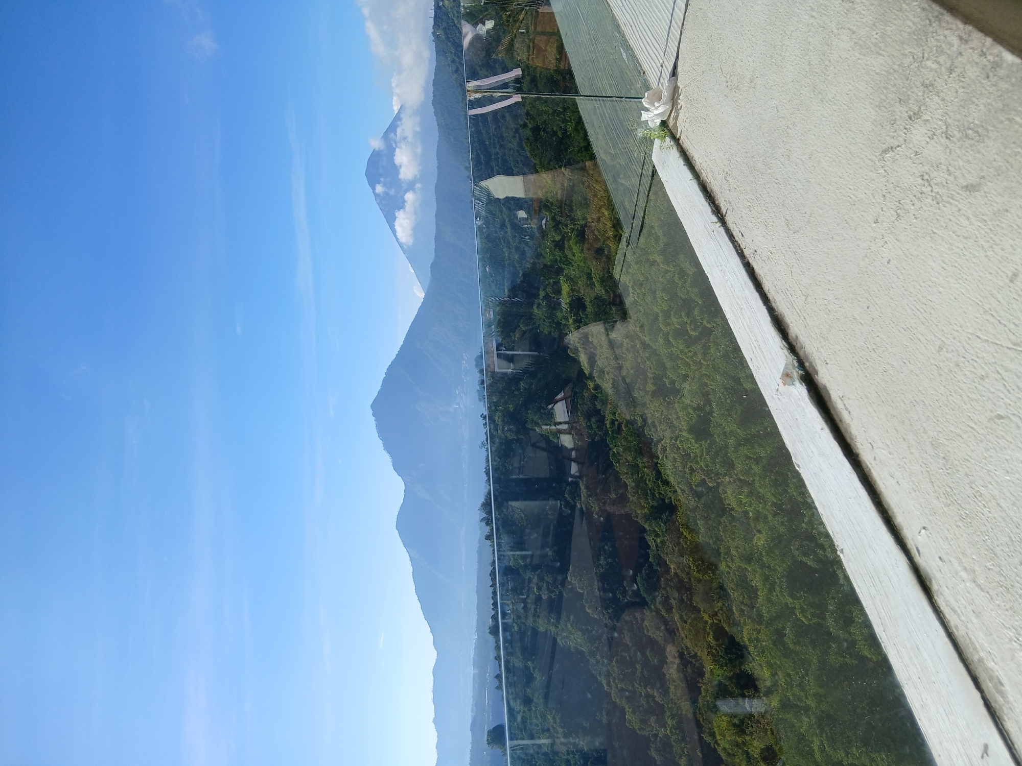 The image captures a scenic view from what appears to be a balcony or elevated vantage point in Kintamani, Indonesia. In the foreground, a concrete structure, possibly a railing or ledge, is visible, with a thin white border and some debris on its surface. A glass barrier extends across the scene, reflecting the lush green vegetation and distant mountains.
Beyond the glass, a verdant landscape unfolds, characterized by dense trees and rolling hills. In the background, a series of majestic mountains dominate the horizon under a clear blue sky dotted with a few wispy clouds. The largest mountain in the distance is partially shrouded in clouds, adding a dramatic effect to the panorama. The overall impression is one of natural beauty and tranquility, typical of the Kintamani region known for its stunning views of Mount Batur and Lake Batur.