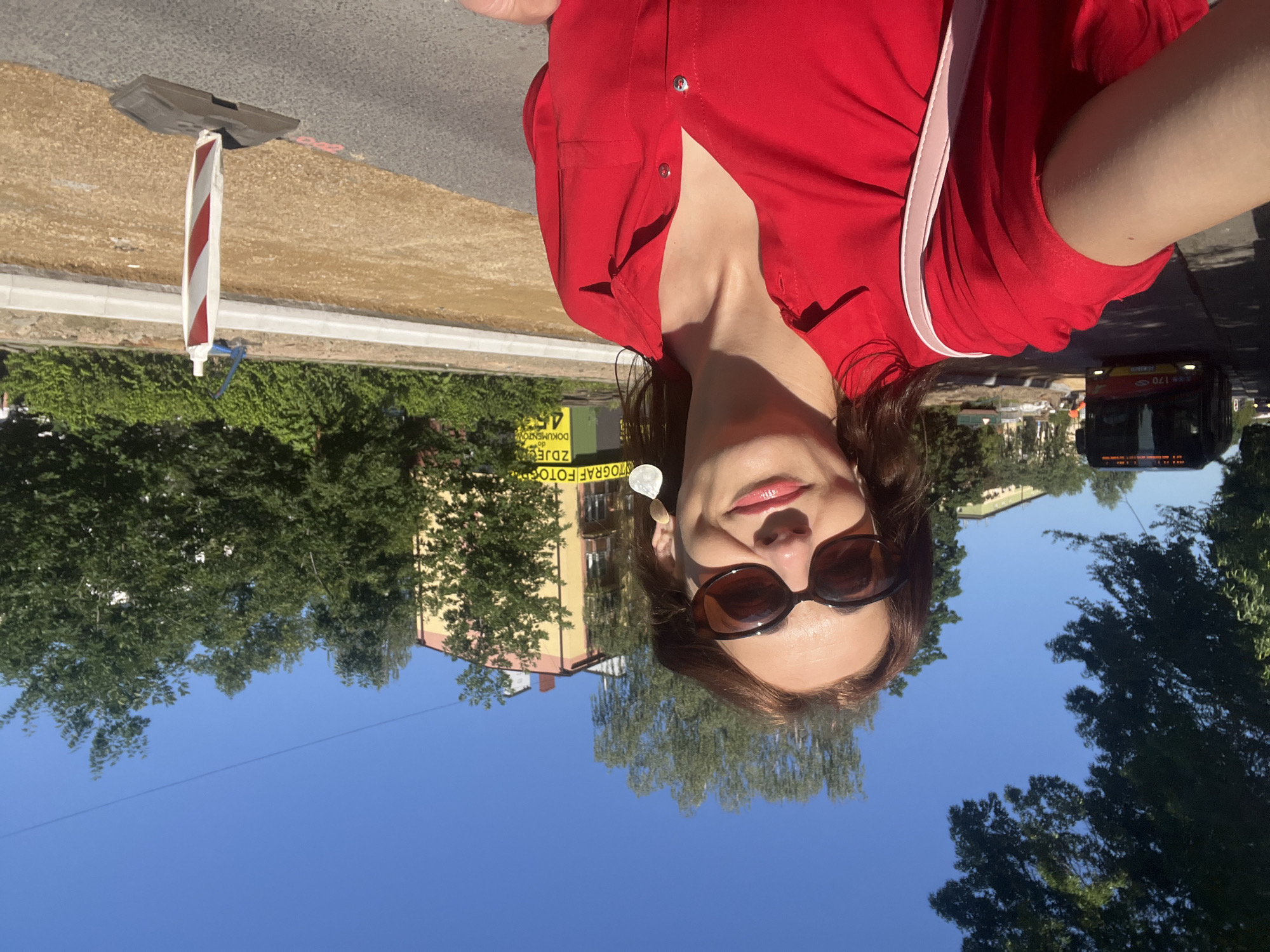 A woman with dark hair and sunglasses is taking a selfie outdoors. She is wearing a red shirt and a light pink strap is visible over her shoulder. The background includes trees, a building, a street, and a bus. A construction barrier is also visible in the foreground. The sky is clear and blue, suggesting it is daytime and the weather is sunny. Text is visible on a yellow banner in the background, partially obscured by trees. The text appears to be in Polish and reads "DOKUMENTACJA FOTOGRAFICZNA" which translates to "Photographic documentation". The overall mood appears casual and relaxed.