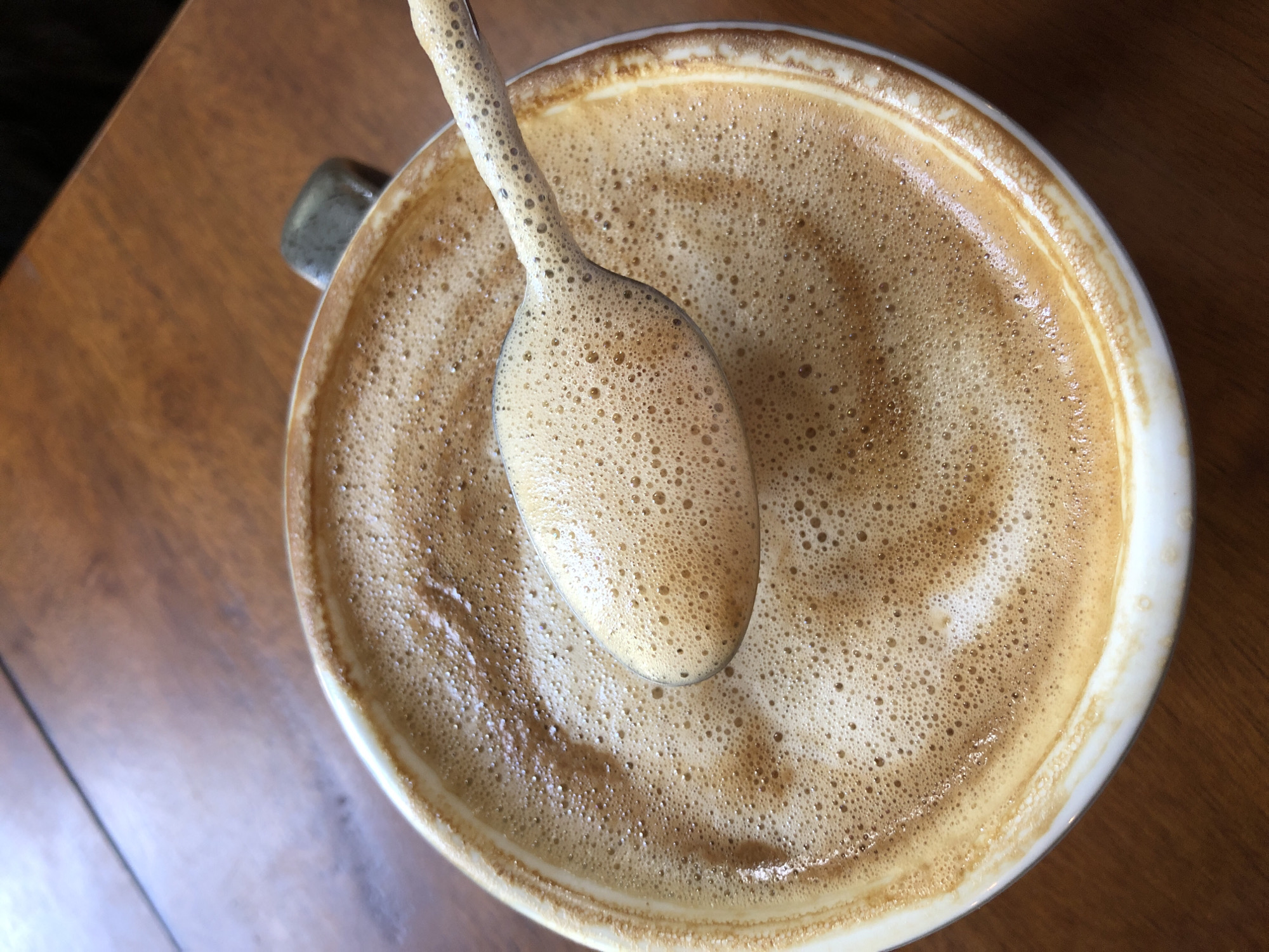 A close-up, overhead shot captures a white ceramic mug filled with frothy coffee, likely a latte or cappuccino, on a dark wooden table. A spoon, also coated in the coffee's foam, rests diagonally across the mug, obscuring some of the beverage's surface. The coffee's surface displays a swirl pattern of lighter and darker browns, indicative of latte art or stirred milk. The mug has a dark gray handle visible on the upper right edge. The overall lighting suggests an indoor setting, possibly a cafe. The image does not depict any specific location in Đà Nẵng, Vietnam, or any individuals.