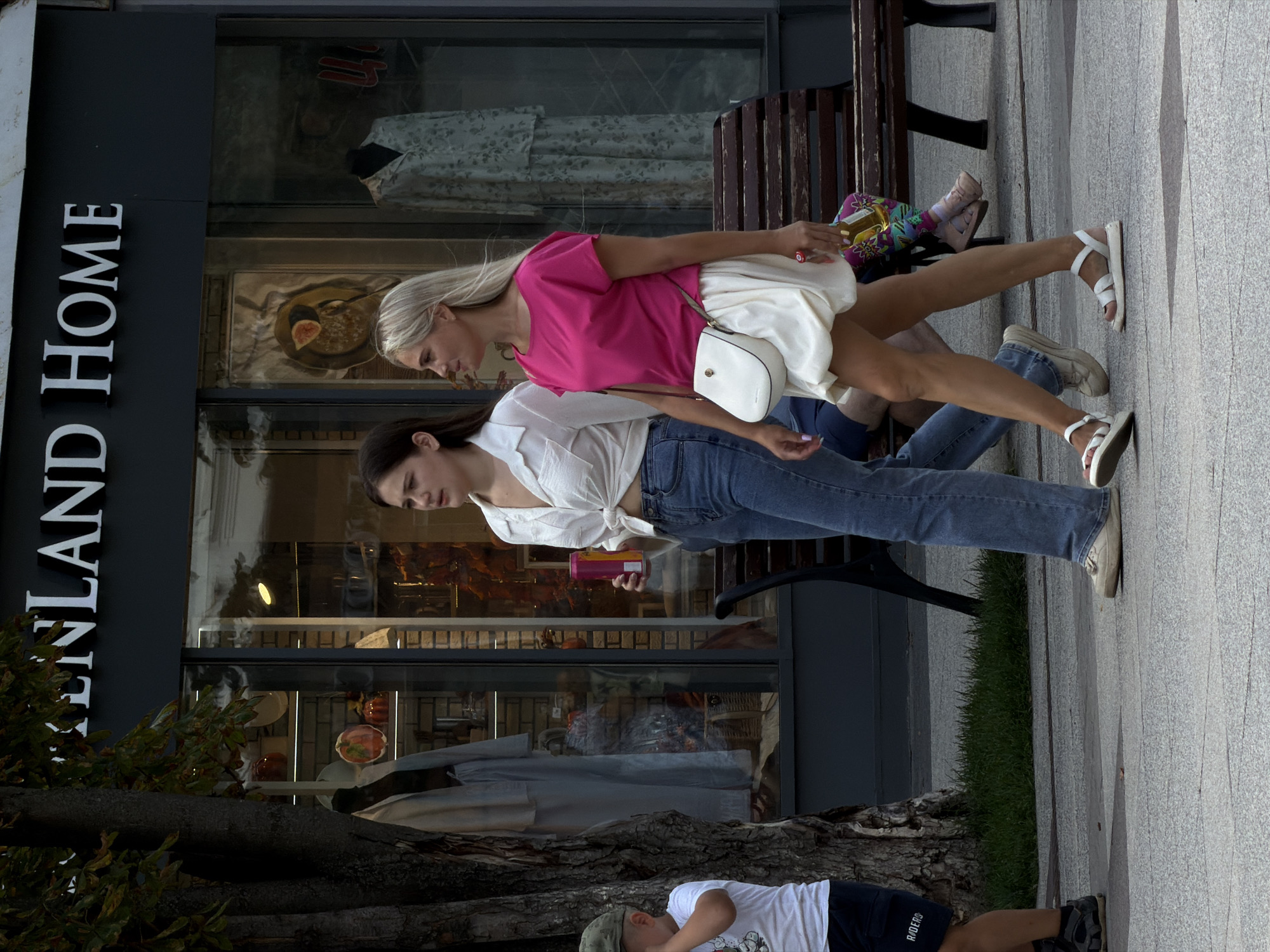 Two women and a child are walking on a sidewalk in front of a store with a sign that reads "LENLAND HOME". The woman on the left is wearing a white, knotted shirt and blue jeans. She is holding a pink and yellow can. The woman on the right is wearing a bright pink top and a white skirt, and carrying a white purse. She is holding a bottle of drink. The child on the left is wearing a green cap and a white t-shirt. The store has large windows displaying merchandise. The sidewalk is made of grey paving stones. A wooden bench is visible behind the women. The scene takes place in Saratov, Russia.