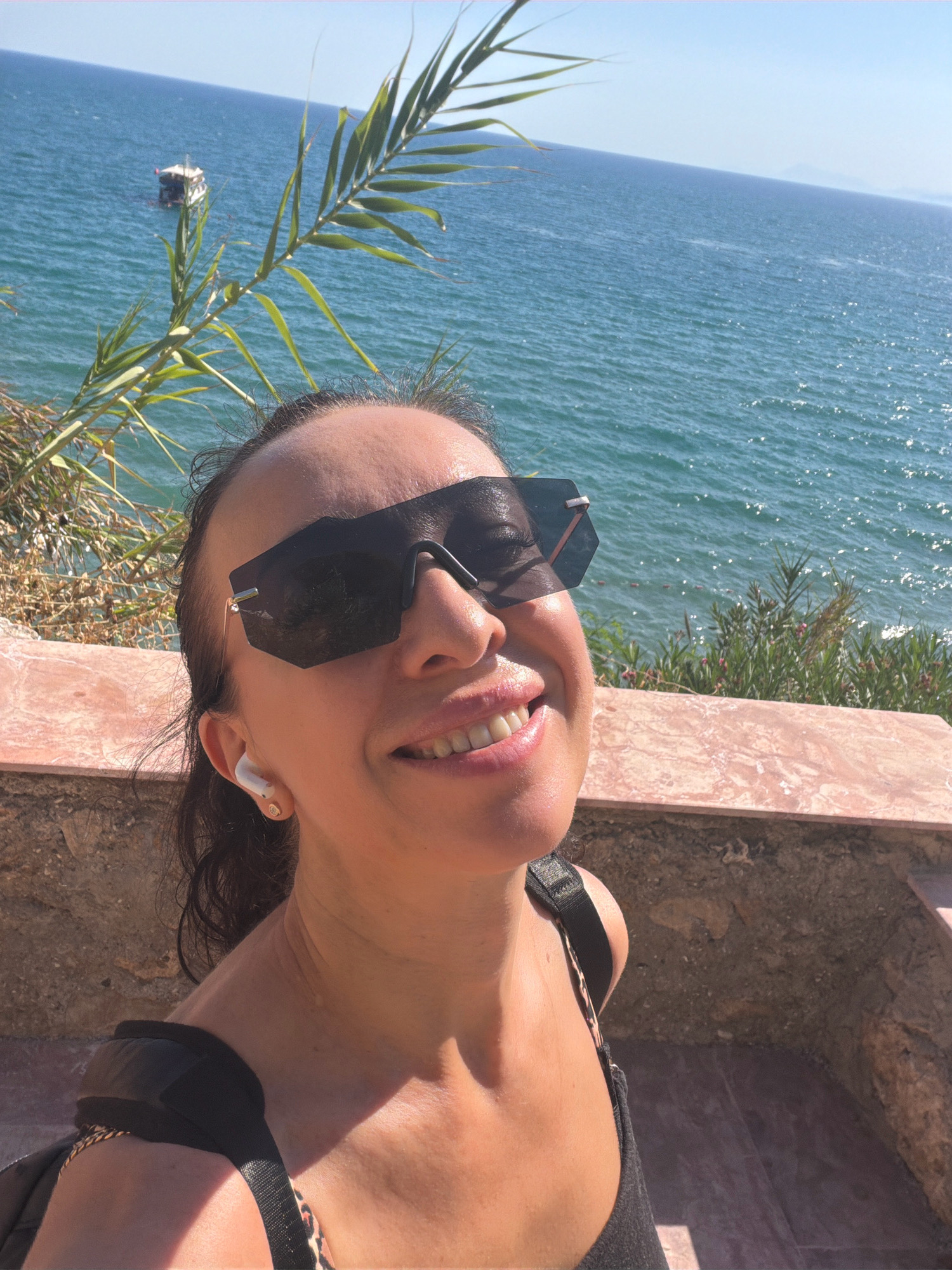 A woman is taking a selfie outdoors, with a body of water and a bright blue sky in the background. She is wearing large, dark sunglasses and has a pleasant expression on her face. Her hair is dark and pulled back. She is wearing a black top and has white earbuds in her ears. A portion of a railing or ledge is visible in the foreground. The water appears to be the Mediterranean Sea, as the image was taken in Antalya, Turkey, a coastal city known for its beaches and clear blue waters. The sunlight suggests it is a warm, clear day. The woman's relaxed posture and smile indicate she is enjoying her surroundings.