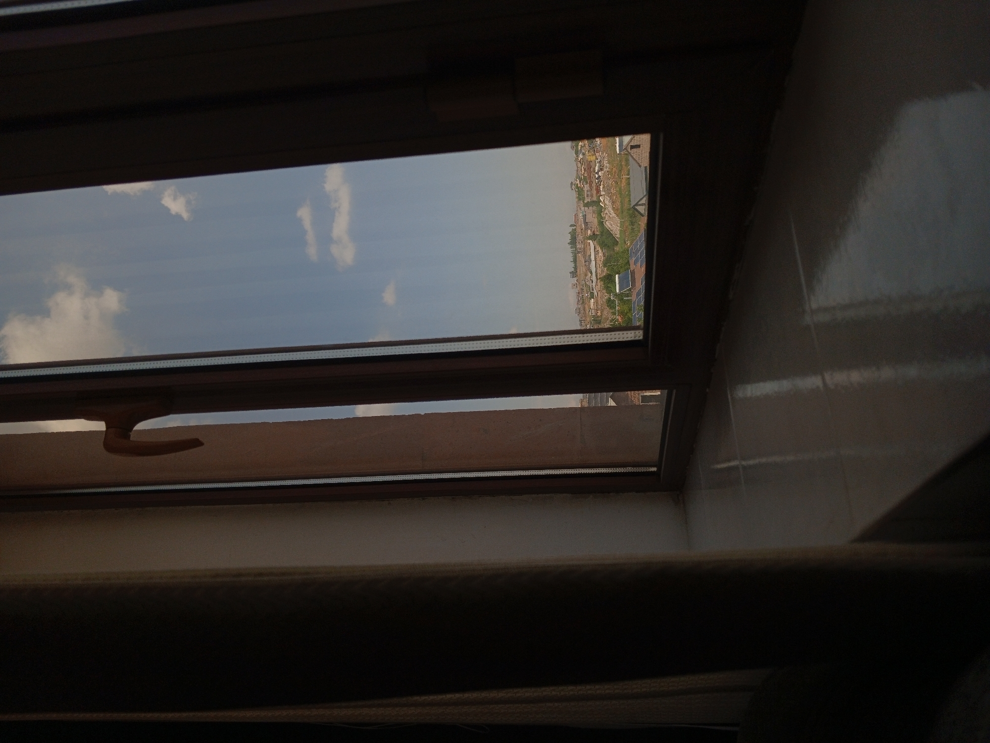 The image depicts a view through a window in Kasakh, Armenia. The window frame is dark brown, with a wooden window handle visible on the right side. To the left of the window, a light-colored curtain is partially drawn, casting a shadow. The windowsill is tiled with a reflective surface, showing glints of light.
The view outside the window reveals a cityscape with scattered buildings, some appearing to be houses with solar panels on their roofs. The architecture suggests a more rural or suburban setting rather than a dense urban environment. The terrain is uneven, with the buildings situated on a gentle slope. Vegetation is visible, indicating an outdoor environment with trees and possibly some greenery. The sky is partly cloudy, with patches of blue sky visible between the white clouds. The overall lighting suggests it is daytime, possibly late afternoon given the angle of the light creating reflections on the windowsill.