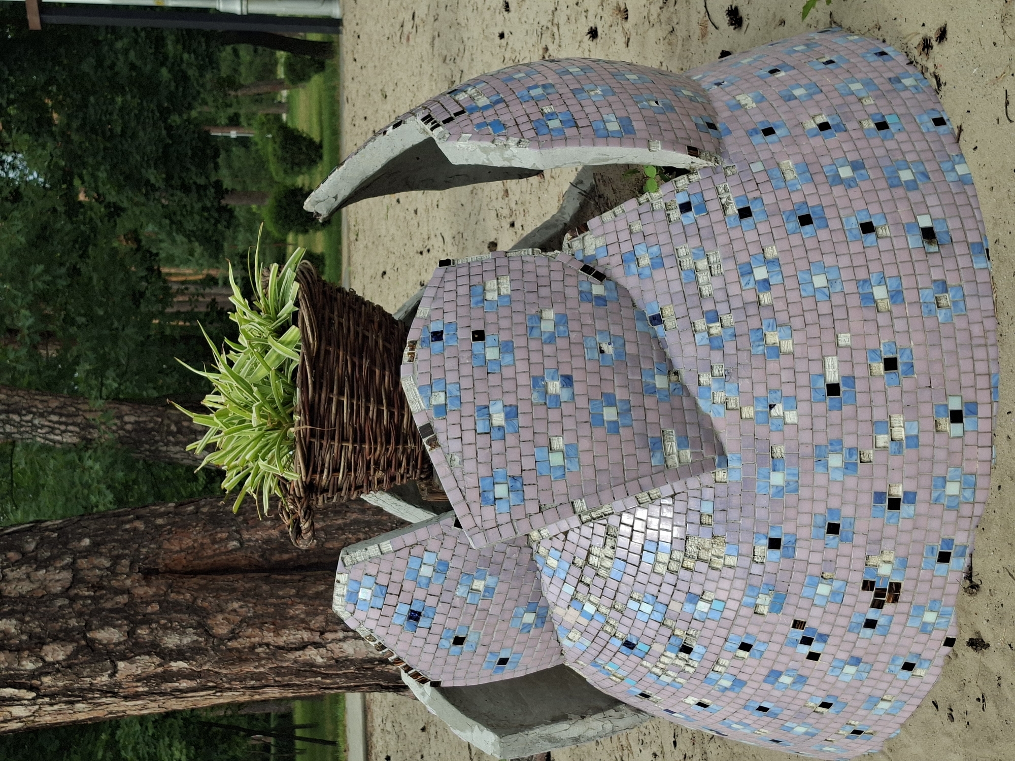 A broken mosaic sculpture is shown in a park setting. The sculpture is made of small, square tiles in shades of pink, light blue, and white, with some black accents. It appears to be in the shape of a large, stylized flower or shell, with several fragmented pieces scattered around. A woven basket filled with green, spiky plants sits within one of the sculpture's openings. The sculpture is placed on sandy ground, with a large tree trunk visible to the left, and a dense green forest in the background. The lighting suggests it is daytime, with overcast or diffused light, indicating it could be a cloudy day or late afternoon. The setting appears to be outdoors in a natural environment, possibly a public park, given the presence of trees and pathways implied by the background. The overall scene conveys a sense of decay and neglect, with the once decorative sculpture now damaged and partially ruined. There is no visible text in the image.