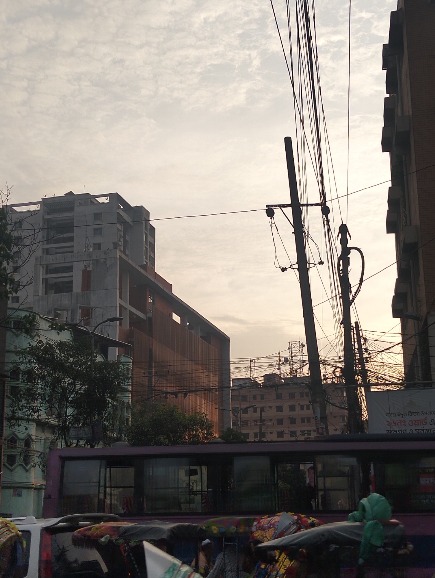 The scene captures a street in Dhaka, Bangladesh. The image is taken at ground level, featuring a bustling urban environment.
Foreground: A partially visible bus is in the foreground. The bus appears to be purple. There are numerous vehicles of unknown type obscuring the scene.
Midground: Power lines and utility poles dominate the midground. Behind them are various buildings, with one appearing to be under construction.
Background: The sky is overcast with clouds.
People: Inconclusive. The bus windows reflect the light, obscuring details within.
Overall: The image portrays a typical, bustling street scene in Dhaka, characterized by its mix of infrastructure, buildings, and heavy electrical wiring.
