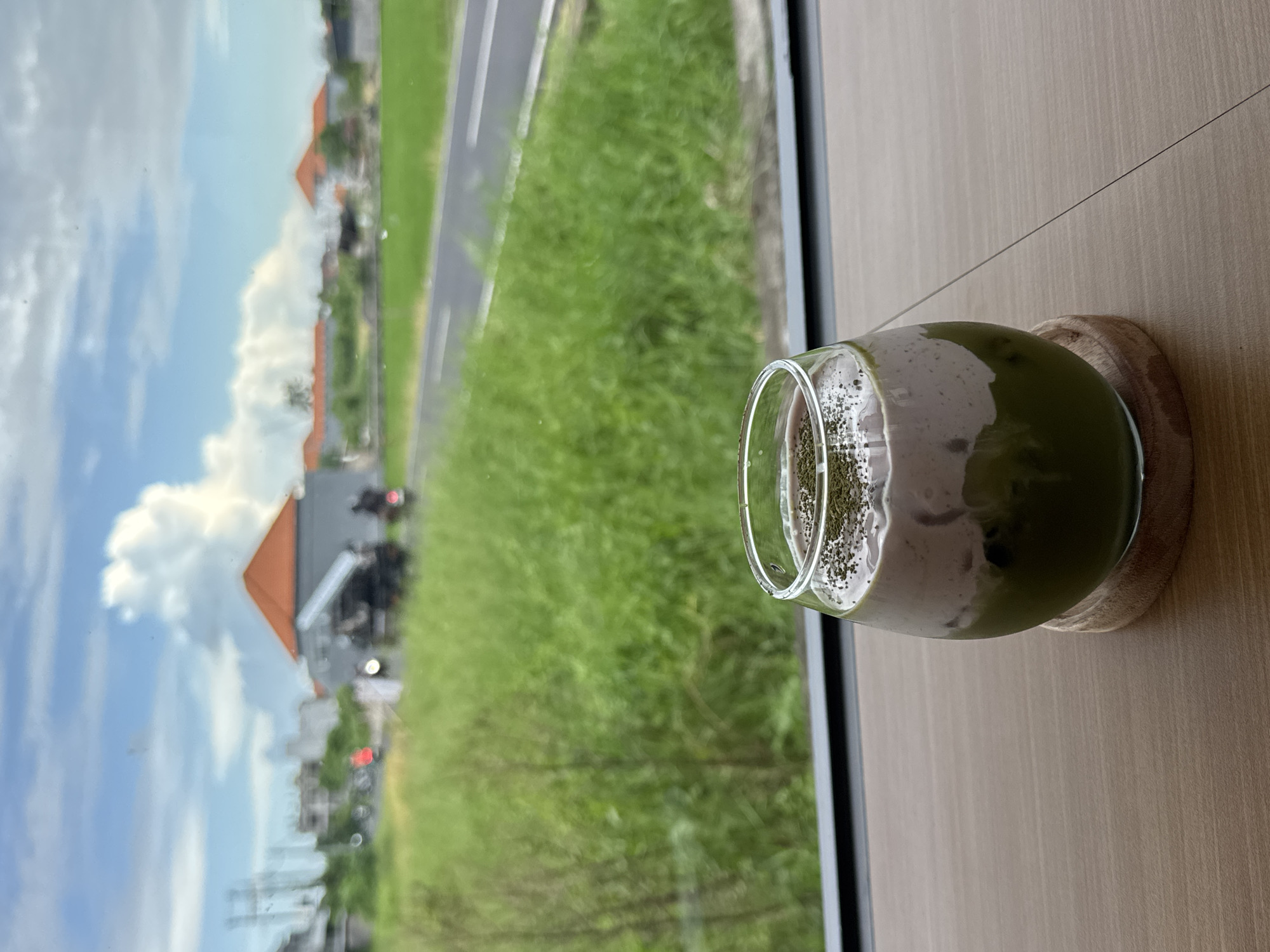A glass containing a layered drink, likely a matcha latte, is prominently featured in the foreground, sitting on a light wood-grained table next to a window. The drink displays a vibrant green layer at the bottom, topped with a creamy, off-white layer, and adorned with dark specks or powder on the surface, possibly chia seeds. The glass rests on a small, circular wooden coaster.
Through the window, a bright daytime landscape is visible. A wide, verdant green field stretches across the midground, with a dark asphalt road or path running along its edge to the left. In the background, under a partly cloudy blue sky, several buildings with distinct orange or terracotta-colored roofs are scattered amongst the foliage. Blurry outlines of what appear to be vehicles or people can be faintly seen on or near the road in the distance. The dark frame of the window borders the outdoor view. The scene evokes a sense of calm and relaxation, set in North Kuta, Indonesia, offering a picturesque view of the local scenery. No text is visible in the image.