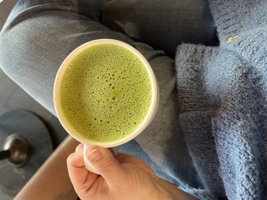 A close-up, overhead perspective shot depicts a person holding a white ceramic mug containing a bright green, frothy beverage. The froth exhibits numerous small bubbles across its surface. The person's left hand, with natural short fingernails, grasps the mug's handle. Visible below the mug are light blue denim jeans on the person's legs, partially covered by a fuzzy, blue knitted garment resting on the lap area. In the blurred background, a dark, cylindrical object, consistent with a table base or chair leg, is partially visible in the upper left, adjacent to a smooth, light brown surface. The scene is indoors. No specific identifying visual markers for the city of Kaiserslautern, Germany are present within the image.