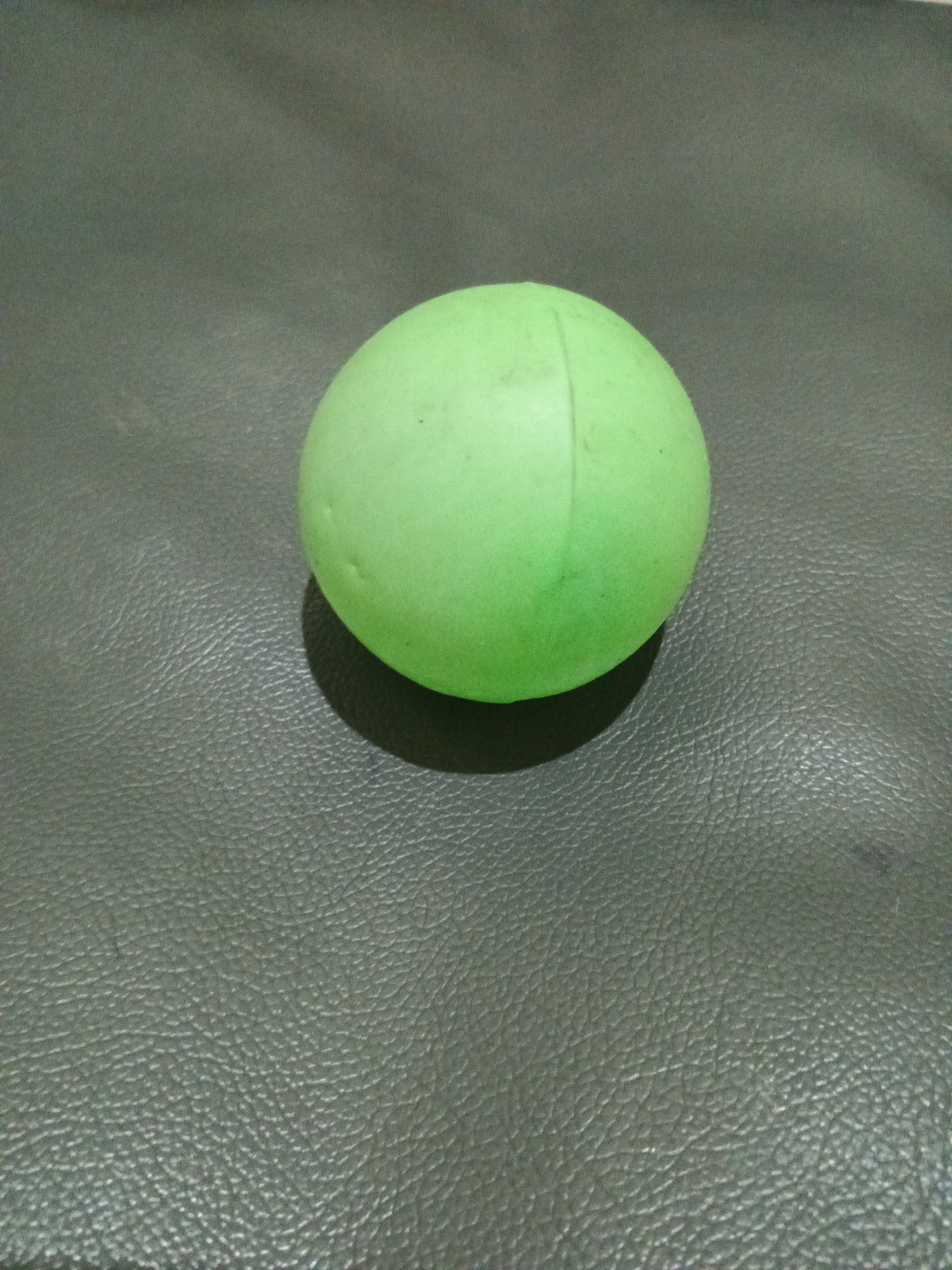 A single, bright green, spherical object rests on a textured, dark surface. The object appears to be a ball, possibly made of rubber or plastic, with a visible seam running across its surface. A faint shadow is cast by the ball onto the dark surface, indicating a light source from the upper left. The dark surface has a grainy texture, resembling leather or a similar material. There are no other objects or people visible in the image. The location is the city of Penambangan, Indonesia.