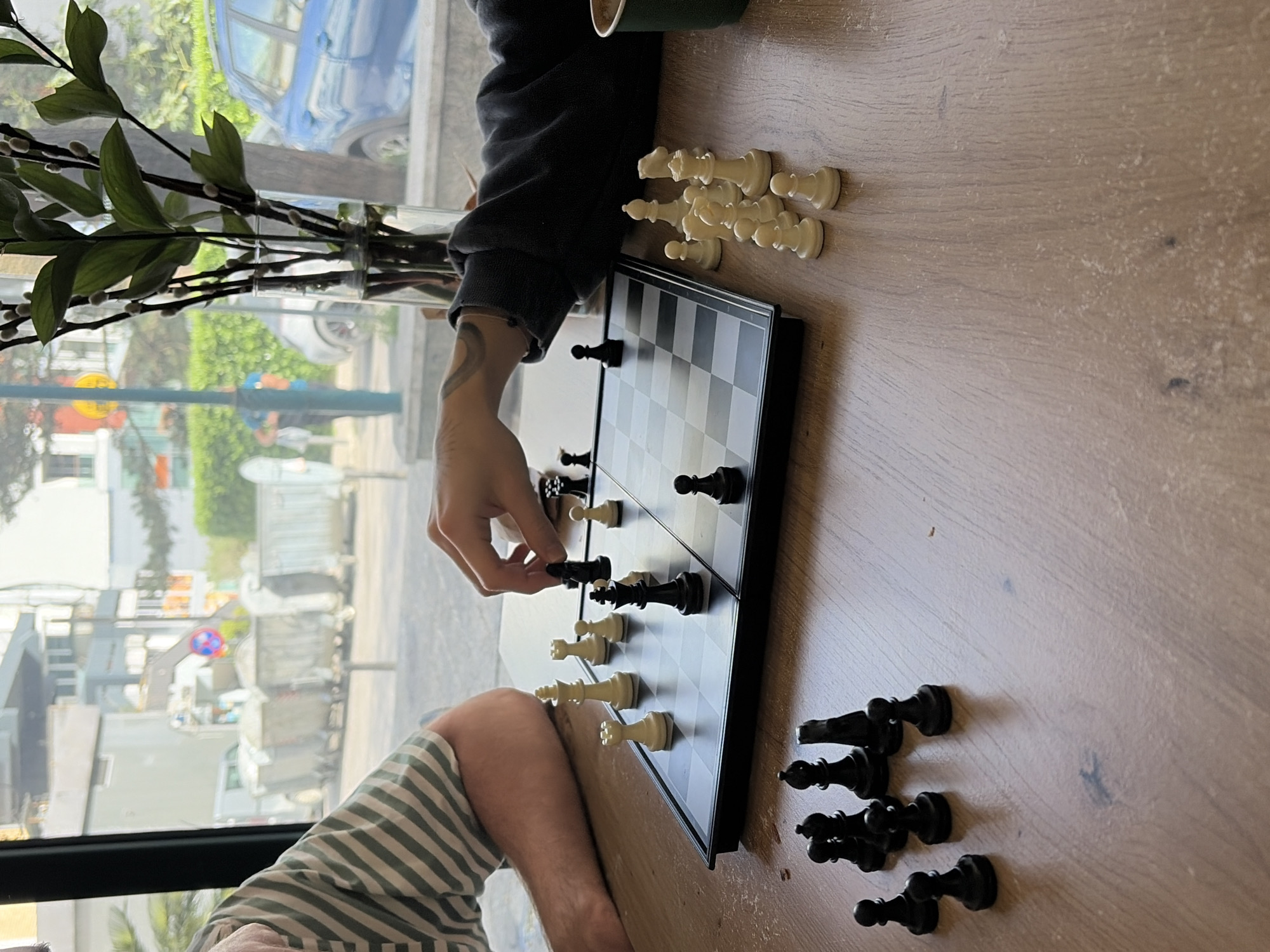 A horizontally oriented medium shot depicts two people playing chess on a wooden table indoors. One person is wearing a black long-sleeved shirt and has a tattooed arm. They are in the act of moving a black chess piece. The other person is wearing a striped shirt. A chessboard is positioned between them, with white chess pieces placed near the person in the black shirt and black chess pieces near the person in the striped shirt. Extra black and white pieces are scattered on the table. A vase of green leaves is visible in the background, along with a window showing a street scene with buildings and a blue car. Based on the architectural style and street layout visible through the window, the location is likely Budva, Montenegro.