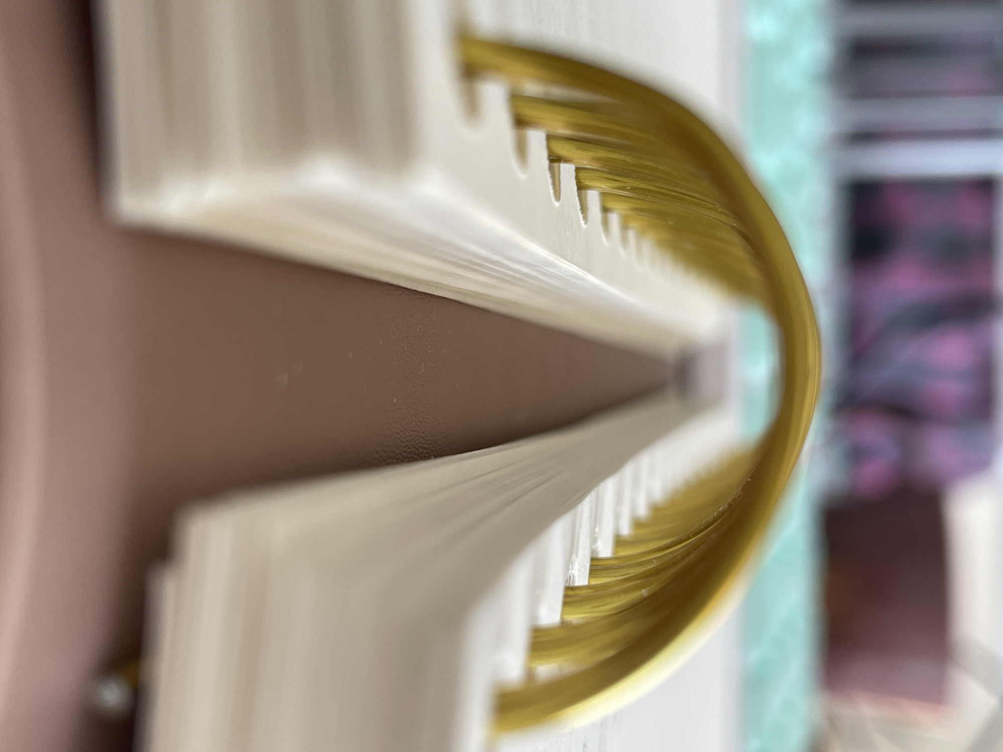 The image presents a close-up, perspective view of a book or journal with golden page edges. The cover is a soft, warm tone, possibly light brown or cream. The pages are trimmed with gold leaf, giving them a shiny, gilded appearance. The binding is a light, creamy white with a slightly textured surface. The background is softly blurred, but appears to contain other objects and perhaps patterned surfaces. The focus is clearly on the book's edge and the detail of the gilded pages. There is no discernible evidence to suggest the location is Izhevsk, Russia, or any other specific geographical place. There are no people or actions visible in the image.