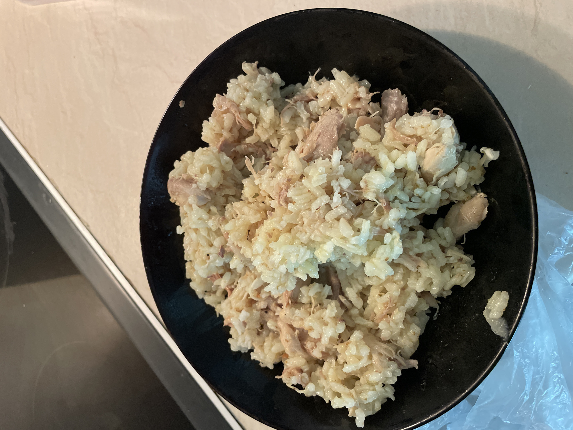 A close-up, overhead shot shows a black bowl filled with a serving of rice and shredded chicken. The rice appears to be cooked and mixed with pieces of white and light brown shredded chicken. The bowl is placed on a light-colored countertop, possibly a kitchen surface. In the background, a dark appliance, likely a stovetop, is visible. The overall impression is of a simple, home-cooked meal. No specific location in Zlatograd, Bulgaria, can be determined from the image.