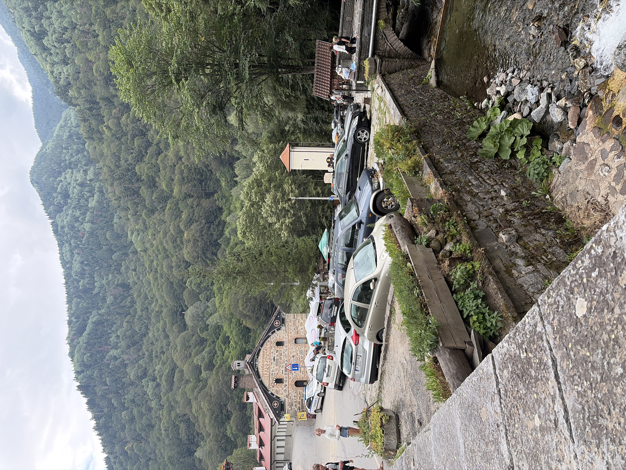 The image depicts an outdoor scene in the Rila Monastery area, Bulgaria. In the foreground, a stone wall and walkway line a flowing stream. To the left, a group of people are gathered near parked cars. A building with a red-tiled roof and stone facade is visible in the midground, with several cars parked in front of it. Further back, a dense forest covers the slopes of mountains under a cloudy sky. A tall, white tower with an orange roof stands near the building. Several vehicles are parked in a lot, including a silver hatchback, a dark SUV, and a black sedan. Some people are walking near the stream and a bridge that crosses it. The environment is natural and mountainous, with a clear stream running through the scene.
