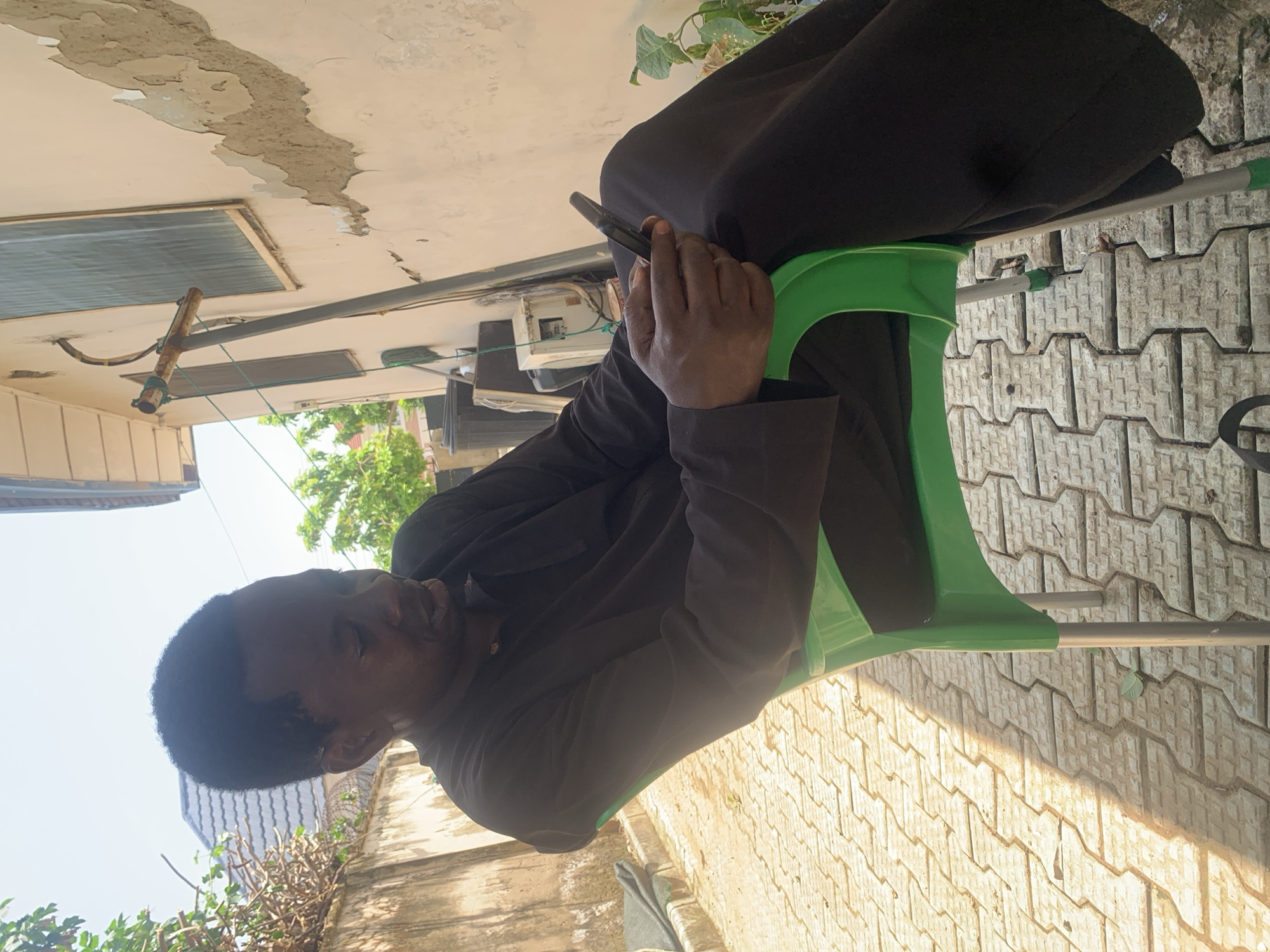 A man of African descent, wearing a dark brown traditional attire, is seated on a green plastic chair with silver metal legs. He is looking down at a black mobile phone he is holding in his right hand. The man has short, dark hair. He is seated outdoors on a paved surface composed of interlocking concrete blocks that are light beige in color. The paved area is partially illuminated by sunlight, casting a distinct shadow across the ground from the man and the chair. In the background, a pale yellow wall with peeling plaster is visible, and to the left of the wall, a section of a building with a dark tiled roof and some foliage can be seen. A metal pole with a curved attachment stands near the wall. The scene appears to be in an outdoor residential or semi-commercial area in Abuja, Nigeria.