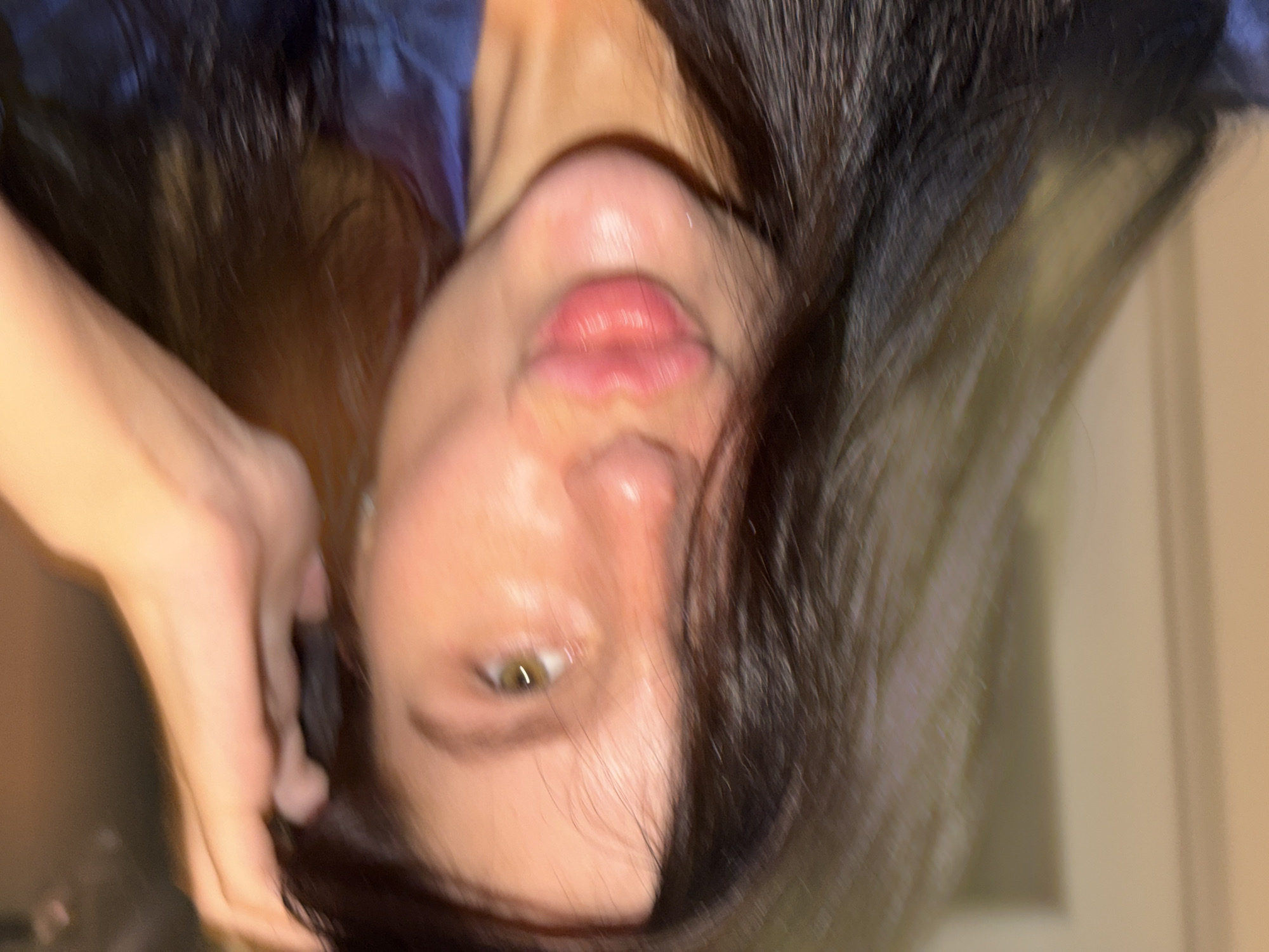 A close-up, vertically oriented portrait captures a person, likely a young woman, with long, dark hair. Her head is tilted, and she looks directly towards the viewer. Her right eye is visible, appearing light green or hazel, and her plump lips are slightly parted or pursed. A hand is seen on the left side of her face, with fingers seemingly running through or adjusting her hair.
Her dark hair frames her face and cascades downwards, appearing somewhat sleek or wet. She is wearing a dark blue garment visible around her shoulders. The setting is indoors, illuminated by warm, artificial light, casting a glow on her skin. The background is blurred, indicating a shallow depth of field, which keeps the focus on the subject's face. The overall image has a soft focus or slight motion blur, contributing to an artistic or intimate feel. No text is visible in the image. The pose and expression convey a sense of contemplation or a carefully composed shot.