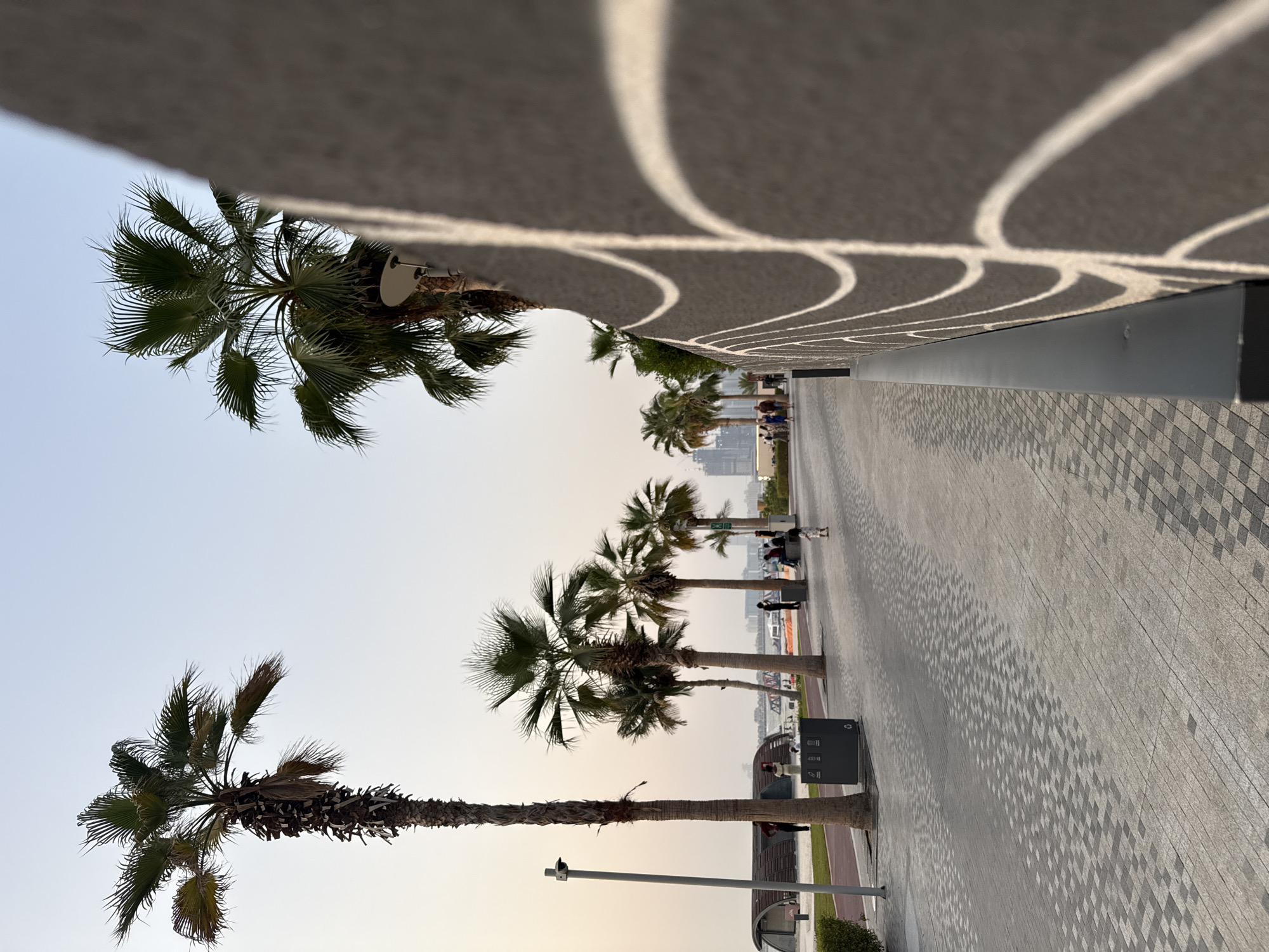 This media file depicts a paved walkway lined with palm trees, likely in Dubai, United Arab Emirates. The scene appears to be outdoors during daylight, with the sun casting distinct shadows of the palm fronds onto the ground, suggesting it might be late afternoon or early morning due to the elongated shadows. The walkway is paved with interlocking rectangular stones arranged in a patterned manner, and further adorned with geometric shadow patterns from an overhead structure.
Several palm trees are visible, with their fronds spread out against a clear, pale sky. In the background, glimpses of modern architecture and distant city structures can be seen, hinting at an urban or developed coastal area. A few people are present, scattered along the walkway, some sitting on benches or walking. One individual is visible with a dog. There are also trash receptacles along the walkway. The overall atmosphere seems serene and leisurely, characteristic of a public promenade or park. The light quality suggests a bright day, but without harsh glare, possibly indicating a slightly hazy or diffused sunlight.