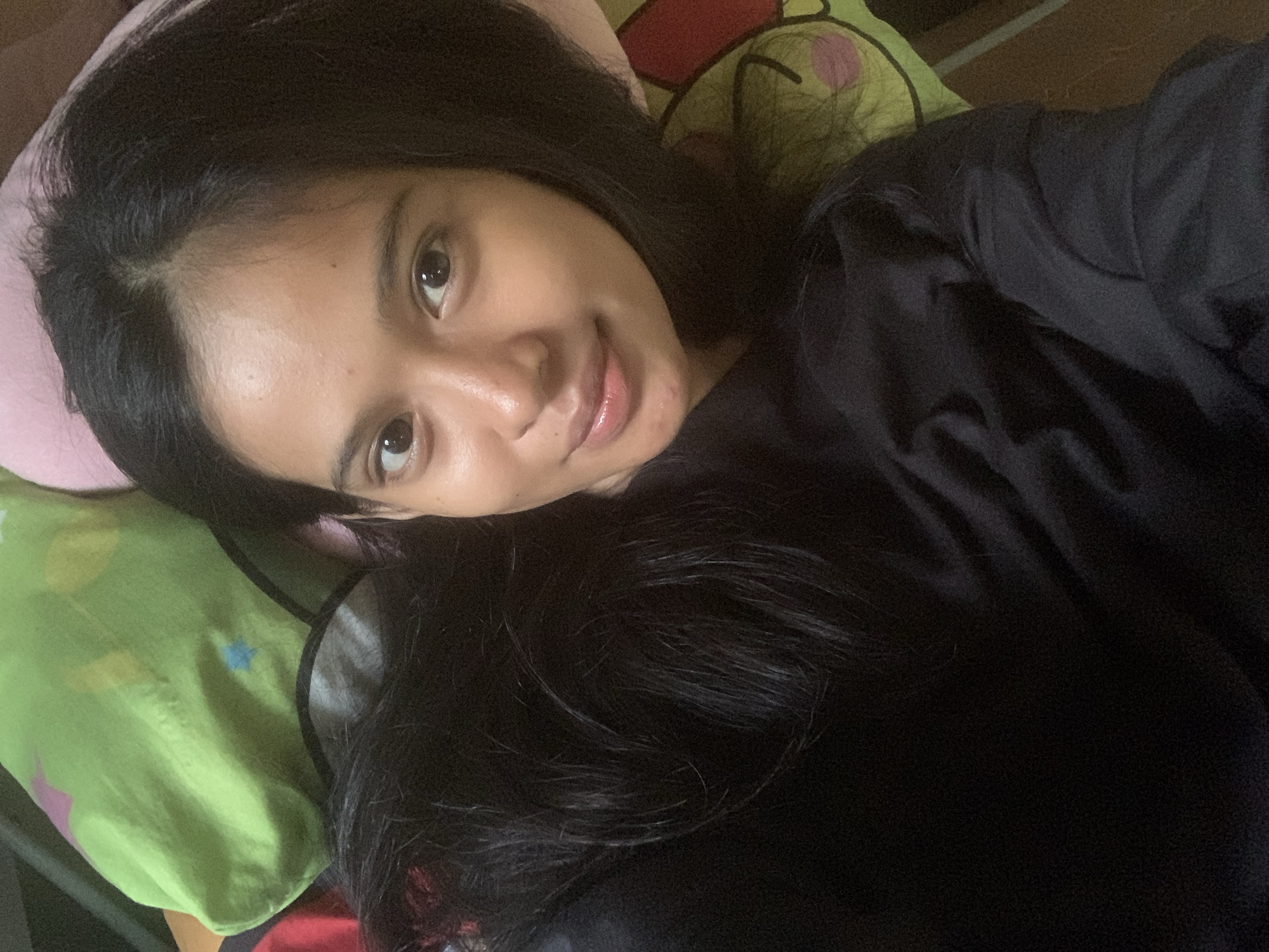 A candid portrait captures a young woman lying down in an indoor setting, likely in Sungai Meriam, Indonesia. She is looking directly at the camera with a soft, relaxed smile. Her long, dark hair is spread around her head and shoulders, partially covering a black long-sleeved top she is wearing. Her skin appears natural and clear.
Her head rests on a light pink pillow, with a vibrant light green pillow visible behind it. This green pillow features playful cartoon-like designs, including a yellow character head with pink cheeks and some blue star shapes. The image is taken from an angled perspective, creating a casual and intimate feel. The lighting is soft and even, suggesting an indoor environment. There is no visible text in the image.