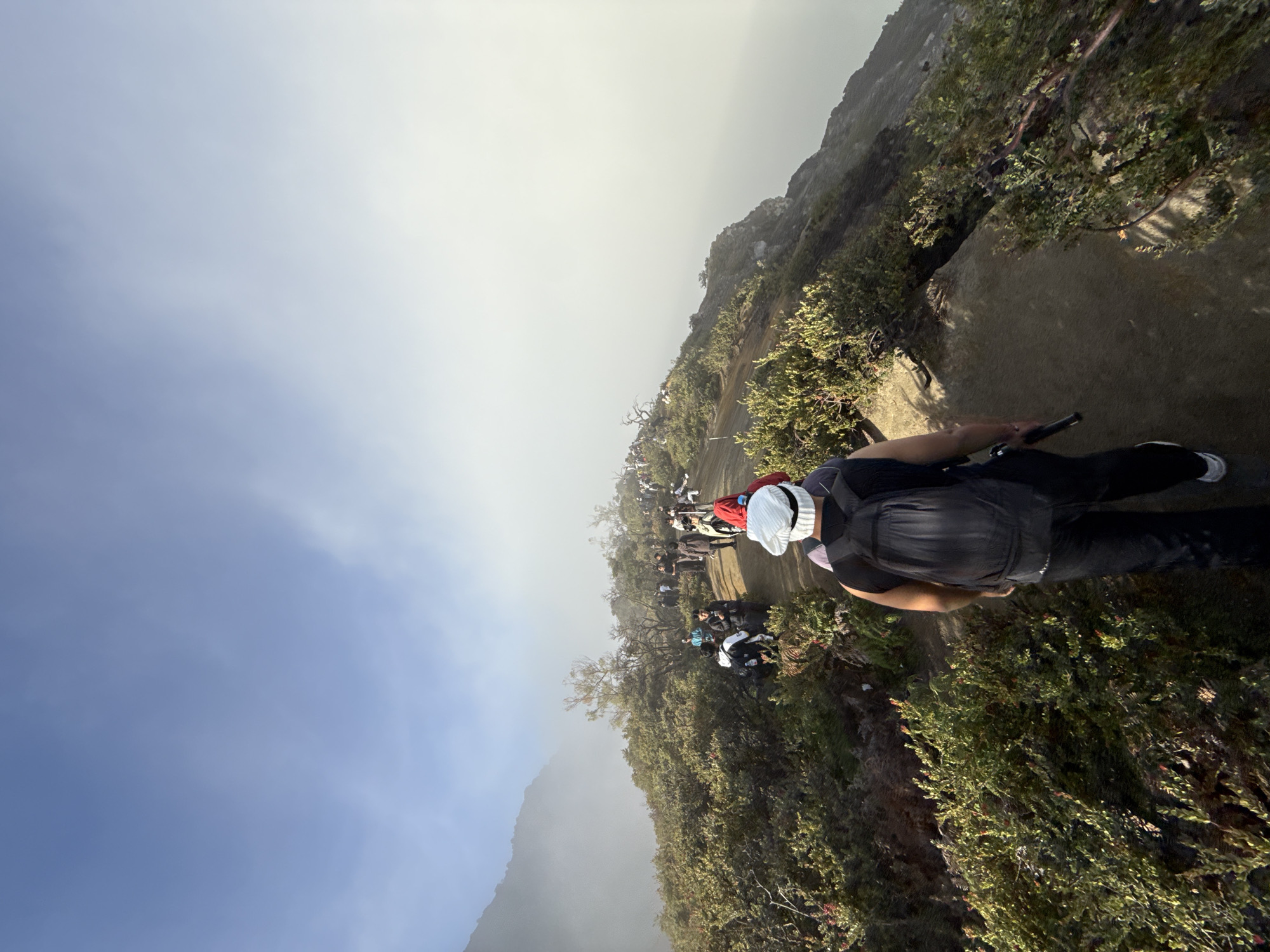 The image depicts a group of people hiking along a narrow mountain trail. The location appears to be Ijen Crater, also known as Kawah Ijen, in Unknown, Indonesia. The sky is partly cloudy with a gradient of light blue to hazy white. The trail is made of dirt or rock and is surrounded by dense green vegetation. A person in the foreground is wearing a dark-colored backpack, black pants, and white sneakers and carrying a device that appears to be a hiking pole or flashlight in their left hand. A group of people are on the trail ahead.