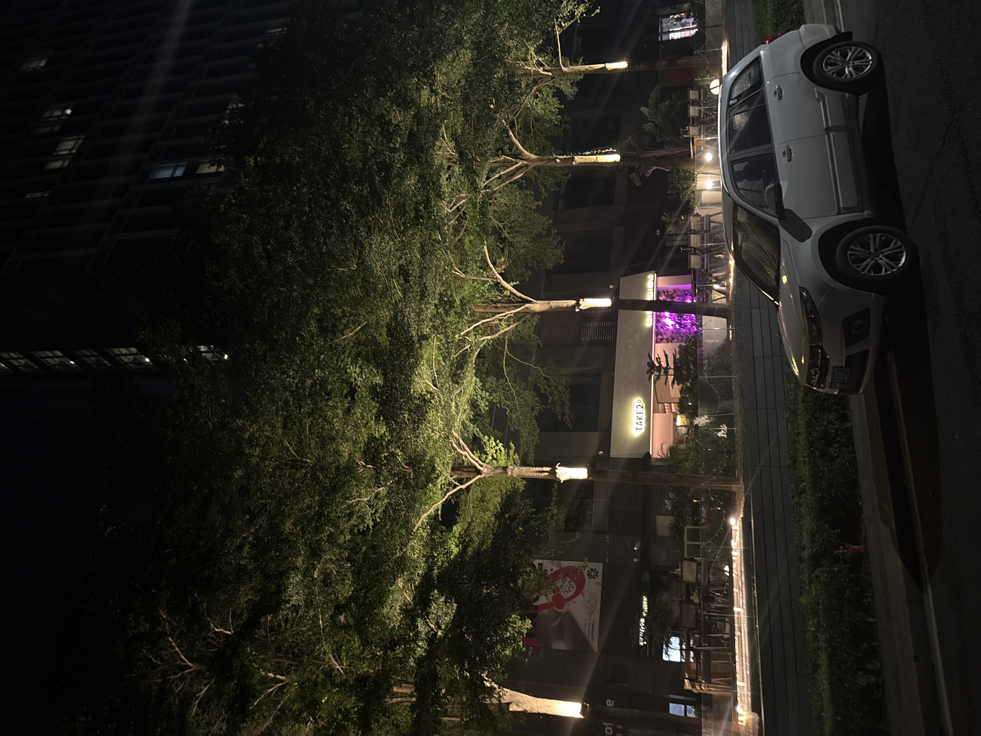 This media file captures a nighttime scene on a city street in Haizhu District, China. The primary subjects are a white car parked on the side of the road and several trees illuminated by artificial lighting. A tall building, appearing to be an apartment complex or office building, dominates the left side of the frame, its windows lit from within, suggesting occupancy. The car is a modern hatchback with alloy wheels. The trees are leafy and well-maintained, with lights placed at their bases, casting an upward glow.
In the background, businesses with outdoor seating are visible, with one establishment displaying purple lighting and another with illuminated signage that reads "TAKE2". Some tables and chairs are set up outside these businesses, indicating a commercial area likely with restaurants or cafes. The overall atmosphere is quiet and urban, with the artificial lighting creating a somewhat dramatic and moody ambiance. The time of day is clearly night, and the weather appears to be clear, with no indication of rain or wind. The image is captured from a slightly elevated perspective, looking down and across the street. There are no visible people in the frame. The scene evokes a sense of urban nightlife and commercial activity.