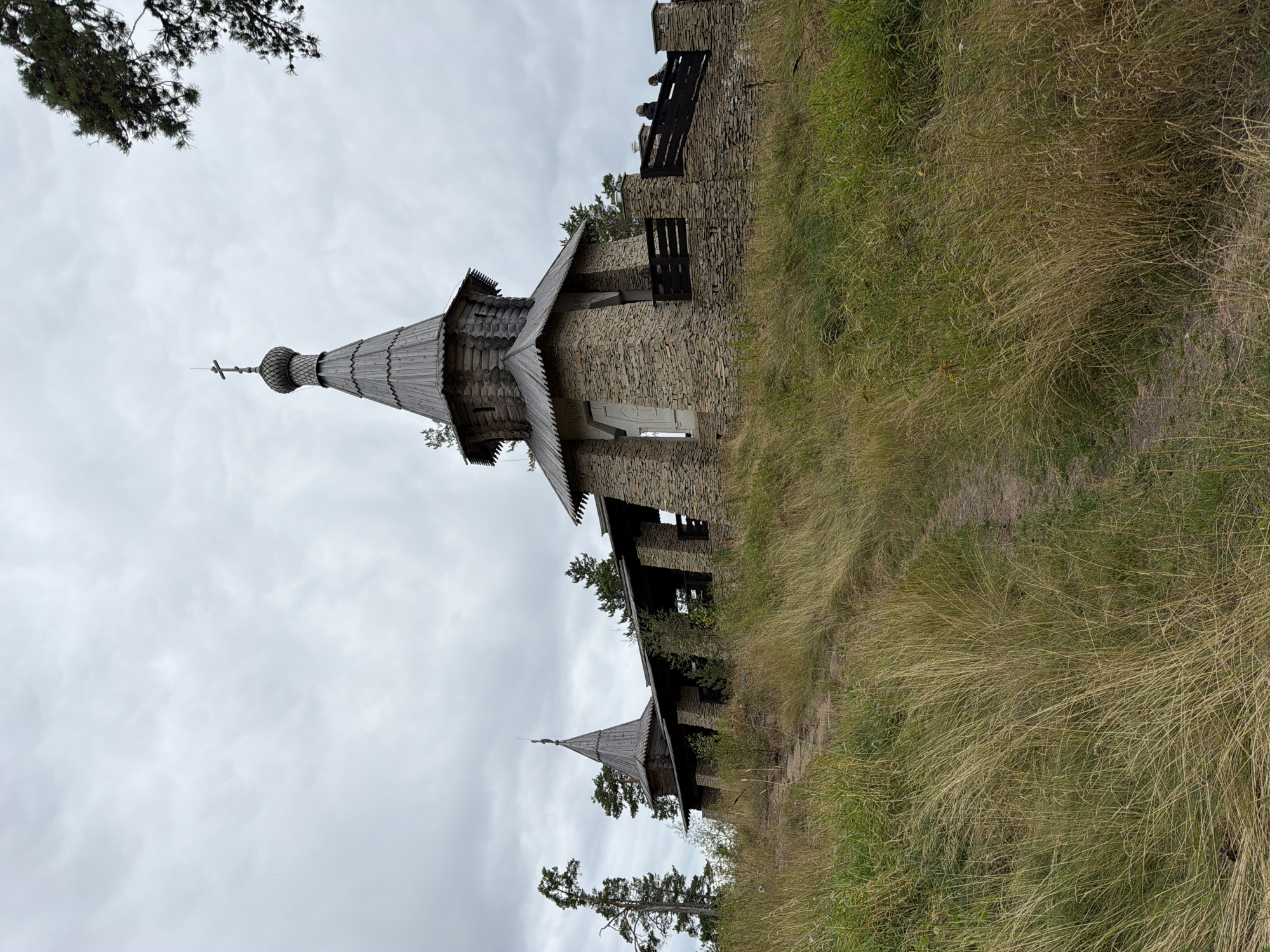The image shows a stone church with wooden elements on a grassy hill. The church has a conical roof topped with a cross and a smaller tower on the left. A stone wall with a wooden railing surrounds part of the church. A dirt path winds up the hill towards the church, through tall, dry grass. Trees, including a tall, thin pine tree, are visible behind and around the church. The sky is overcast with gray clouds. Three people are visible on the walkway behind the wall on the right side of the church.
