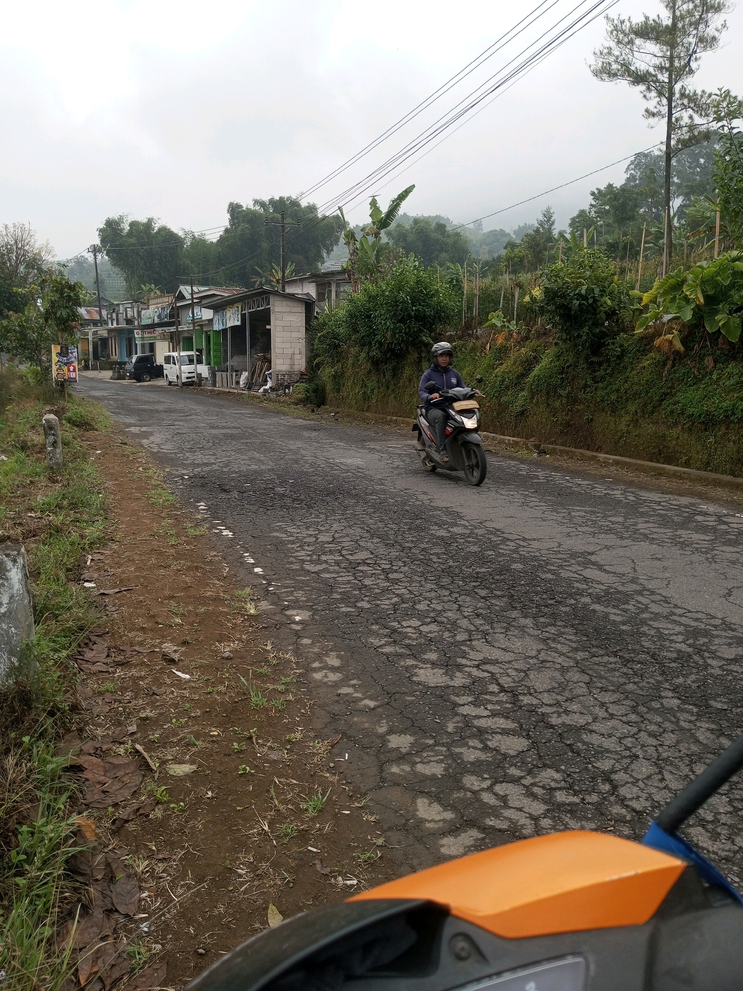 In Keseneng, Indonesia, a man rides a dark-colored scooter along a cracked asphalt road. The scooter has a dark blue jacket and a white helmet. The road is lined with small businesses on the left and a lush, green hillside on the right. The sky is overcast, and a light mist hangs in the air, obscuring the distant mountains. The scene suggests a rural or semi-rural setting, possibly in a mountainous region.