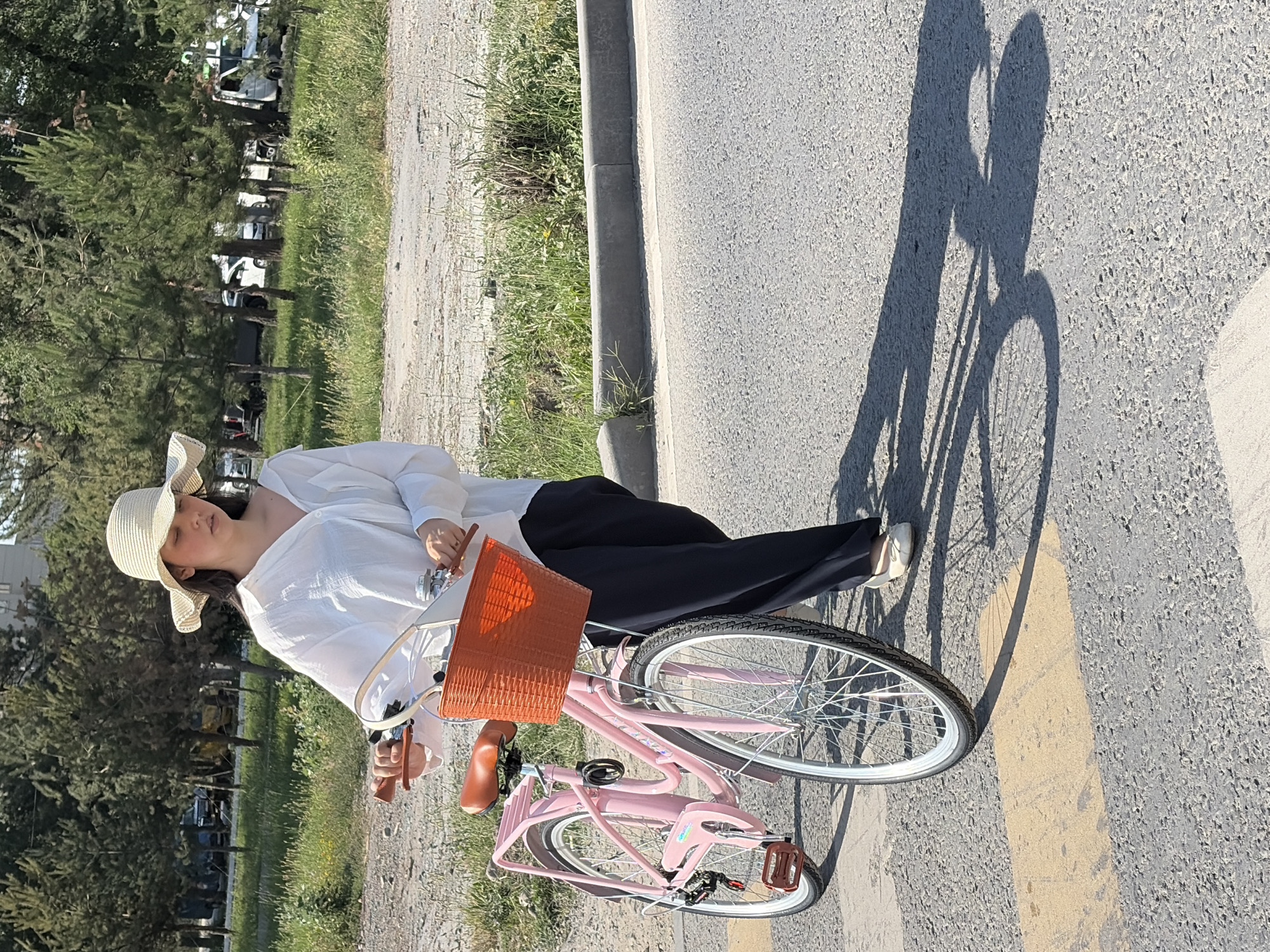 A woman in a white shirt, dark pants, and a wide-brimmed hat pushes a pink bicycle with an orange basket on a street. The bicycle has a brown seat and pedals. The woman is walking on a crosswalk with yellow markings. In the background, there are trees, grass, a curb, and several parked vehicles. The scene appears to be outdoors in a city.