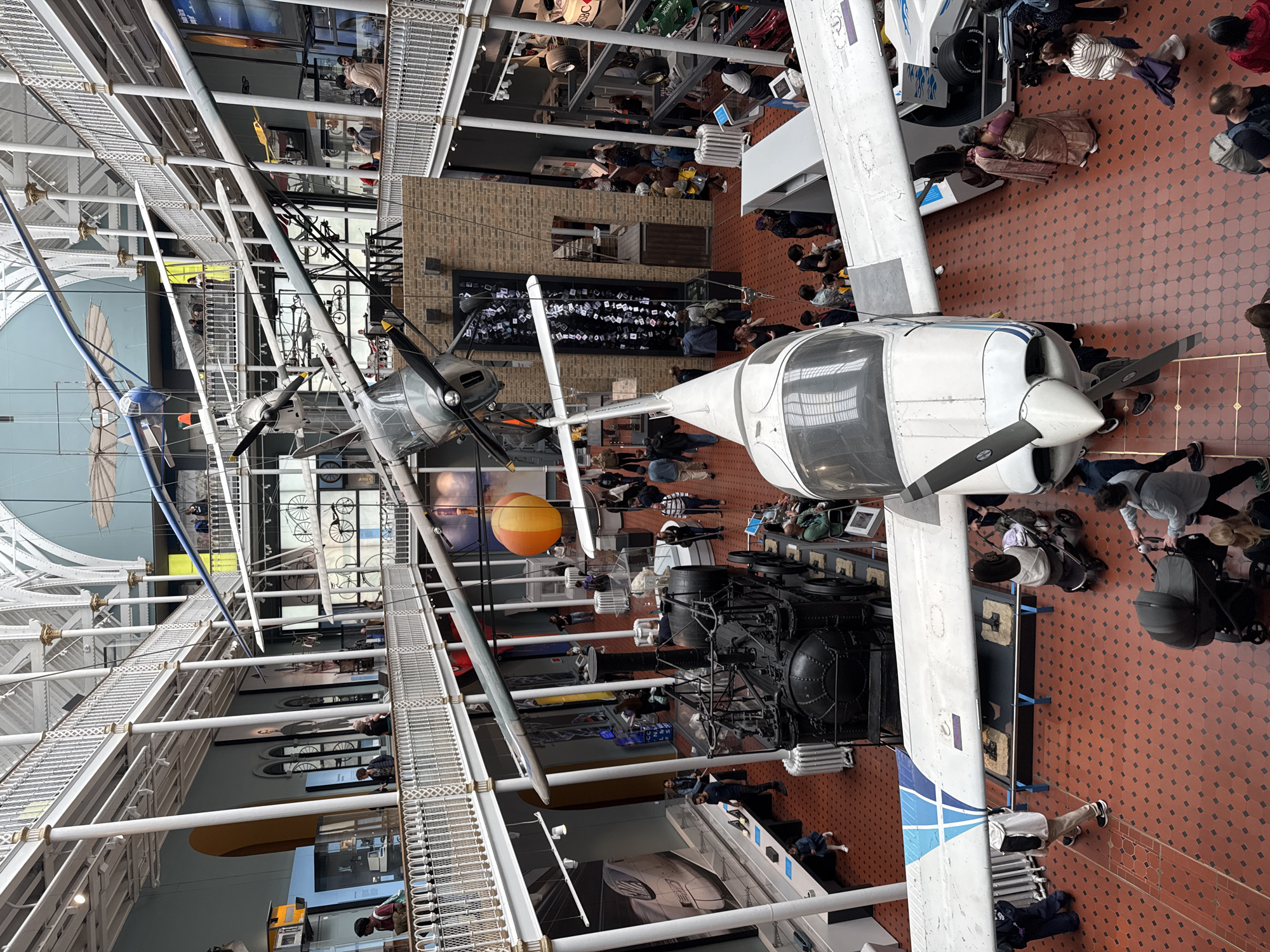 The image shows the interior of the National Museum of Scotland in Edinburgh, United Kingdom. The museum features a large atrium with multiple levels of balconies overlooking a central exhibition space. Various modes of transportation are displayed, including several aircraft suspended from the ceiling, a vintage steam engine, and a Formula 1 race car. People are seen browsing the exhibits, walking on the balconies, and interacting with each other. The floor is tiled with red and black geometric patterns, and the architecture of the building is visible, with white balconies and exposed metal beams supporting the roof. The overall scene depicts a busy museum environment with a focus on historical and technological artifacts.